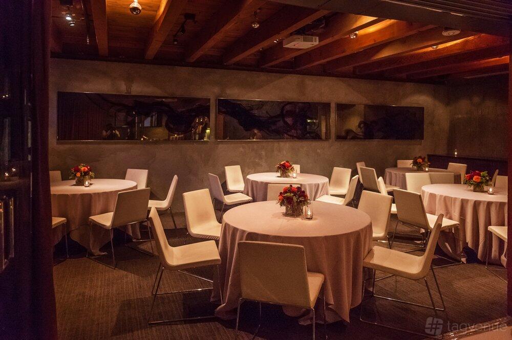 A dining room with round tables draped in pale linens, white chairs, and exposed wood ceiling at 25 Lusk.