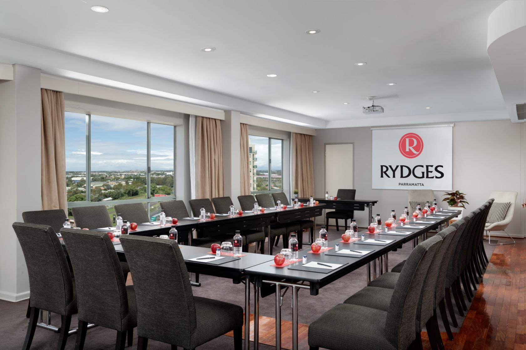 A function room with U-shaped tables, black chairs, large windows, and city views at Rydges Parramatta.