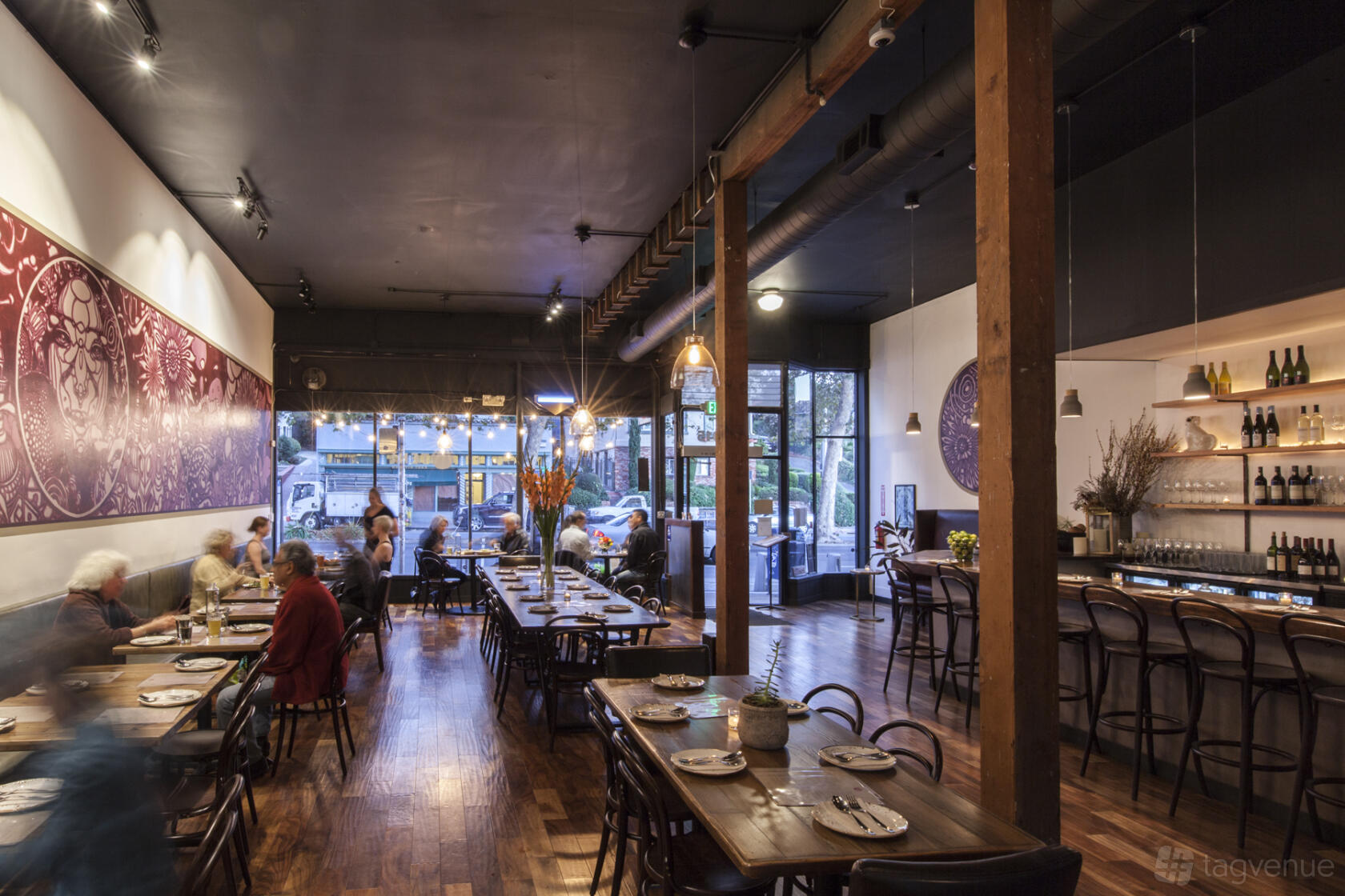 A restaurant dining area with wooden floors, long communal tables, and a bar with bottles at Teni East Kitchen.