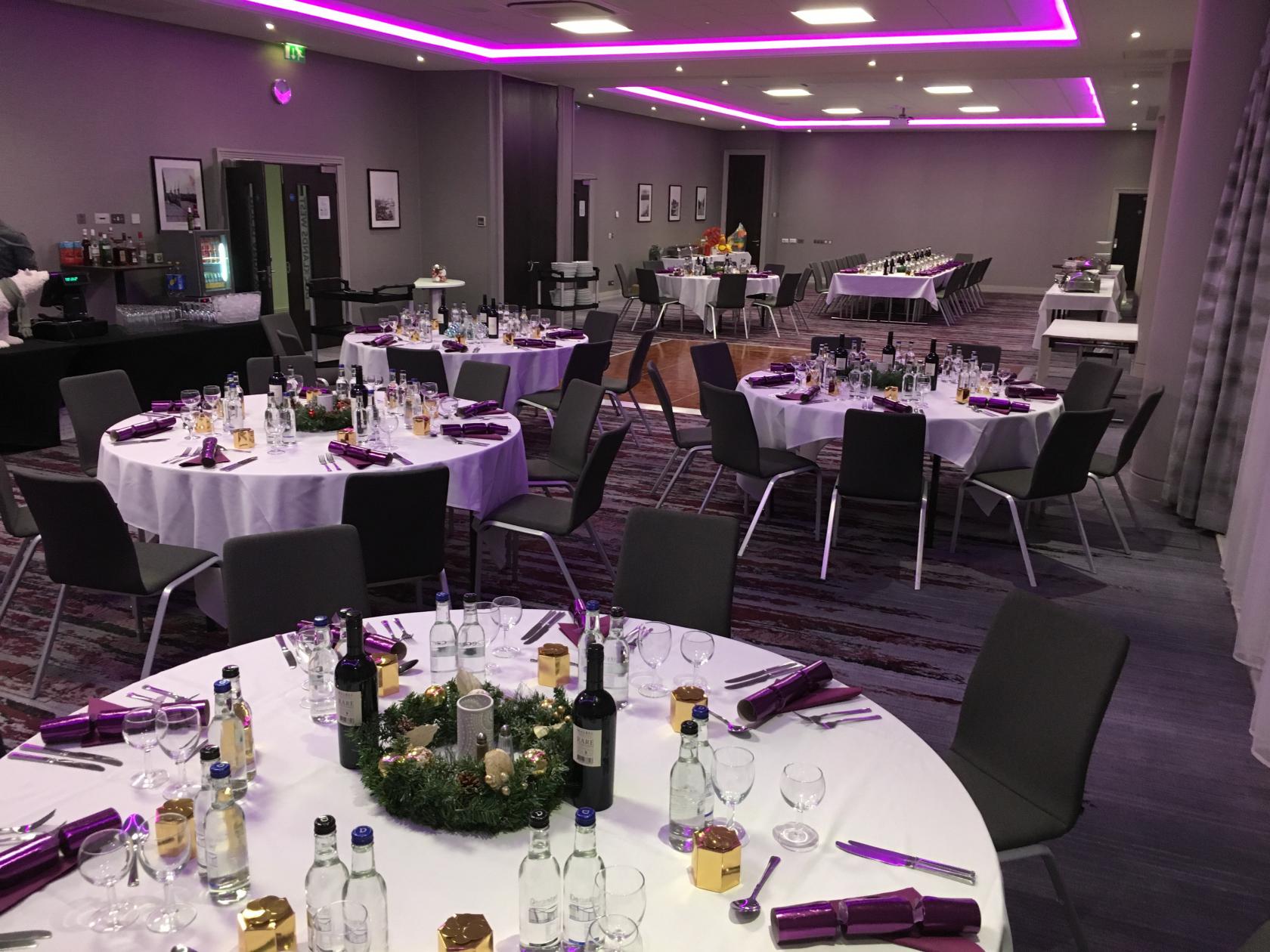 An event space with round tables covered in white linens, banquet chairs, and purple accent lighting at Crowne Plaza London Docklands.