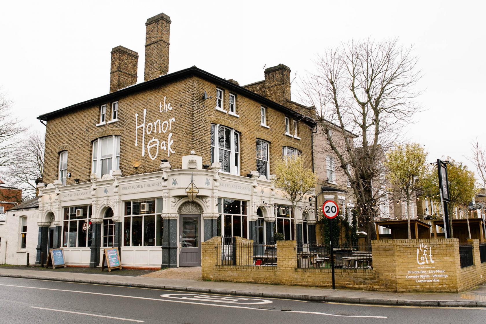 A brick pub with large windows, outdoor seating area, and white painted signage at The Honor Oak.