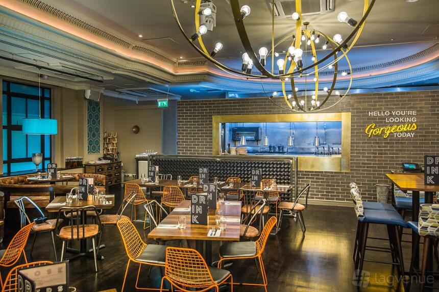 An event space in a bar with a mix of wood tables, colorful wire chairs, an open kitchen, and decorative lighting at Sway.