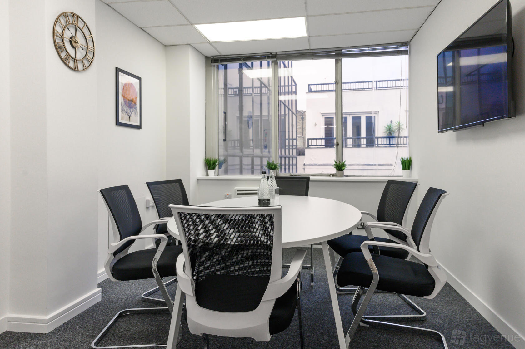 A meeting room with a round table, six mesh chairs, large windows, and a wall-mounted TV at Easyhub Chelsea.