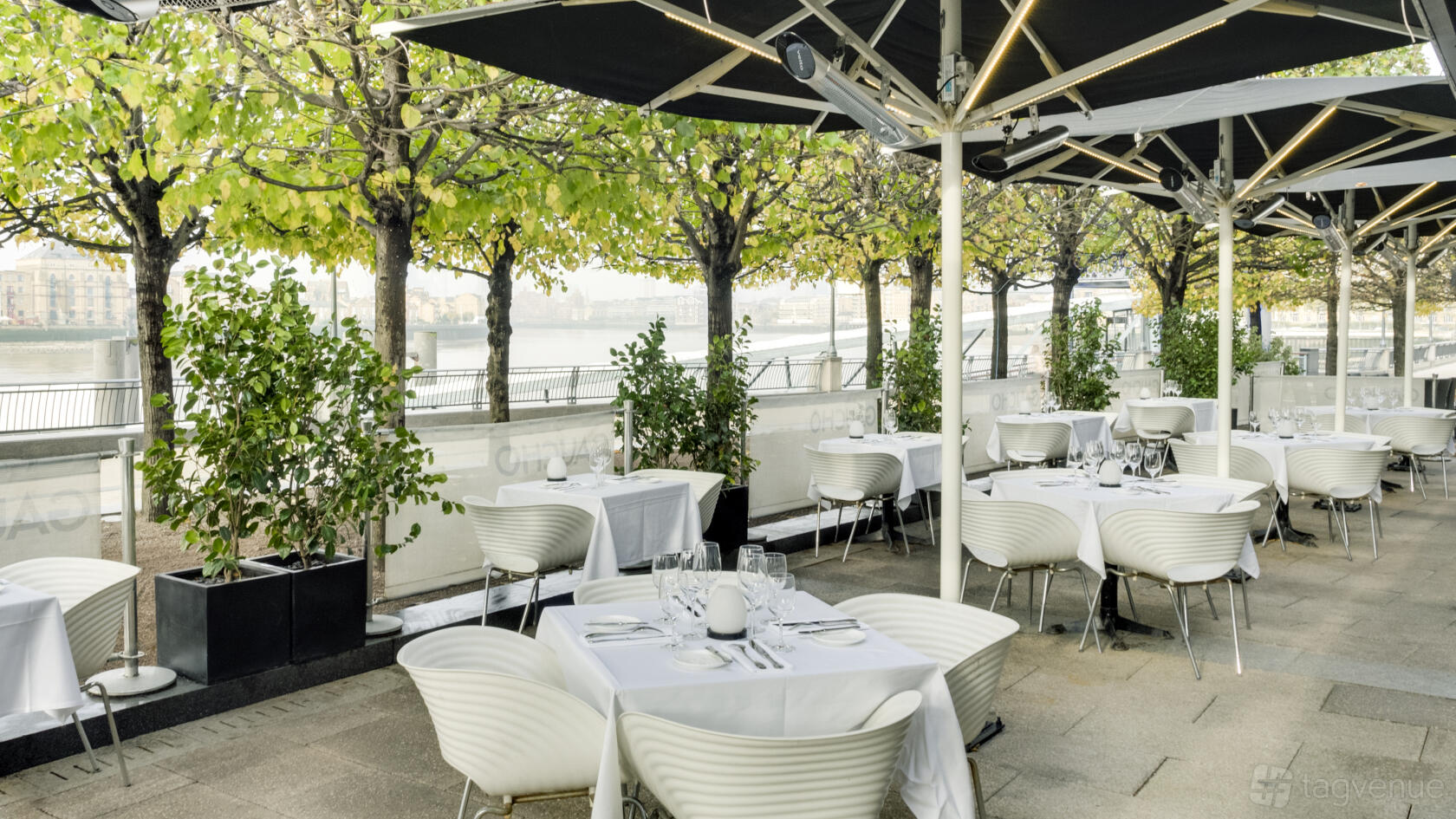 A restaurant terrace with white tables and chairs, canopy covering, and leafy trees at Gaucho Canary Wharf.