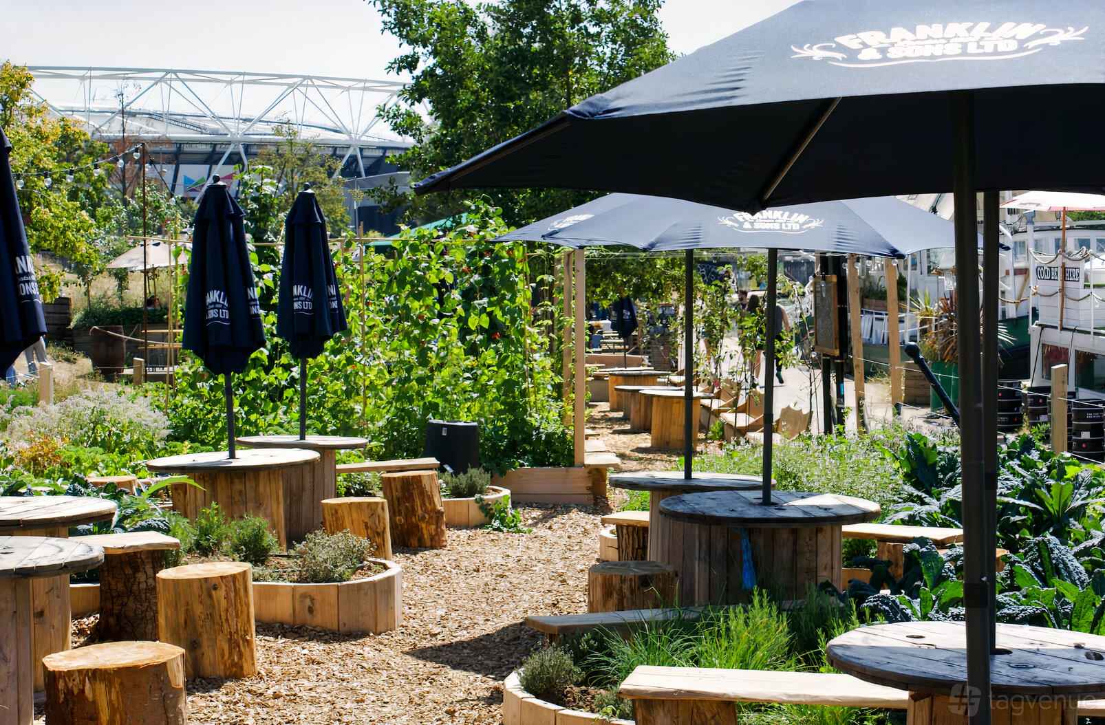 An outdoor restaurant garden with round wooden tables, log seating, and large umbrellas at Barge East.