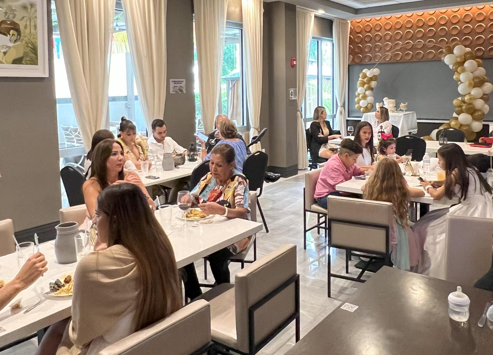 A dining room with marble-top tables, upholstered chairs, and large windows with sheer curtains at Tabbouleh Grill.