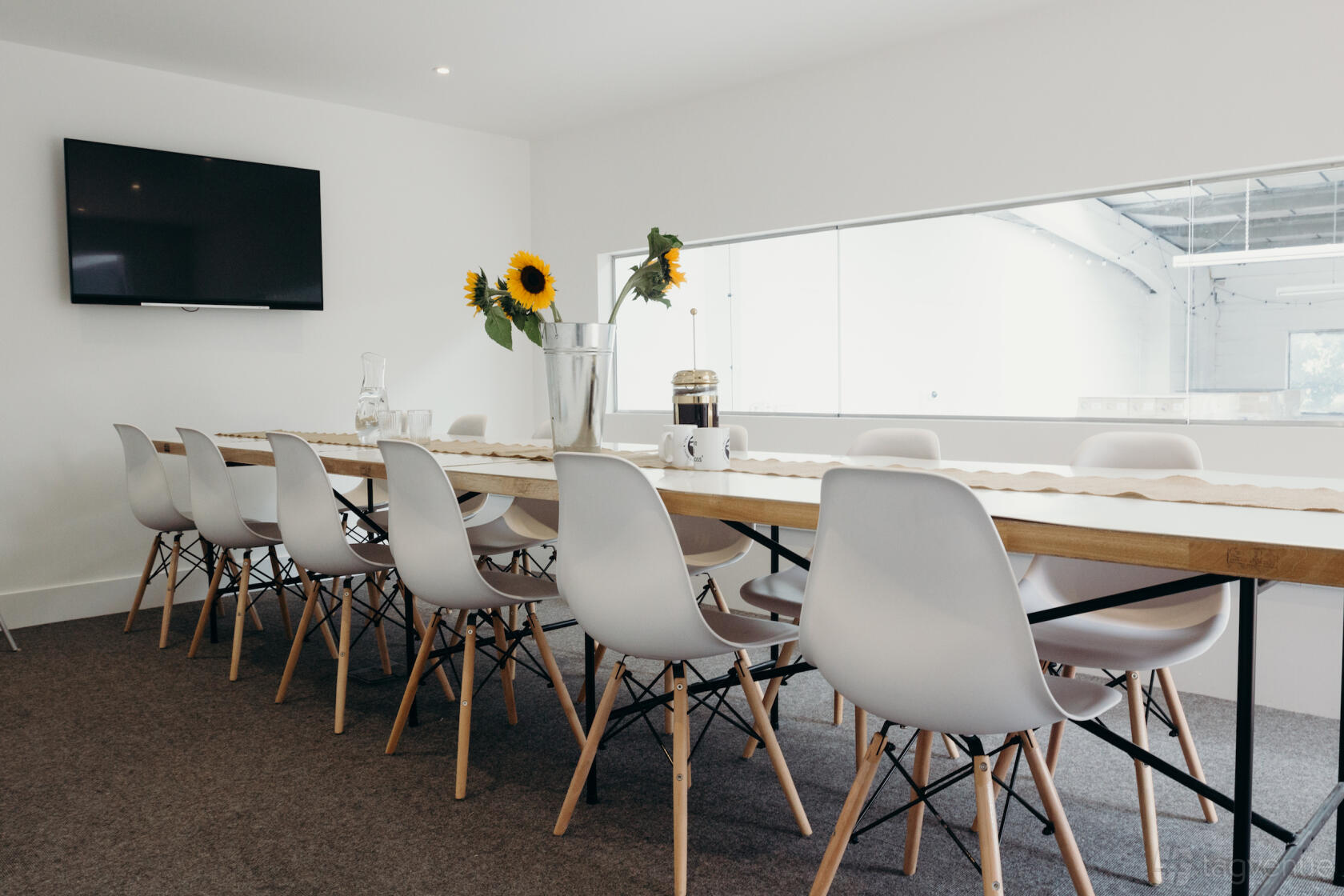 A boardroom with a long wooden table, white chairs, sunflowers in a vase, and a wall-mounted TV at Small Beer Brew Co.