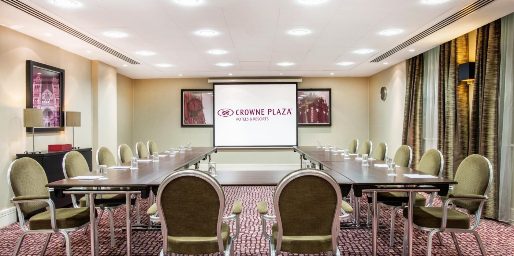 A meeting room with U-shaped tables, green chairs, carpeted floor, and a projector screen at Crowne Plaza London Ealing.
