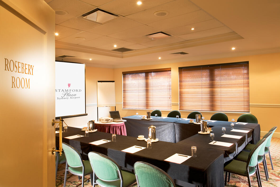 A hotel meeting room with U-shaped tables, green chairs, and a projector screen at Stamford Plaza Sydney Airport Hotel.