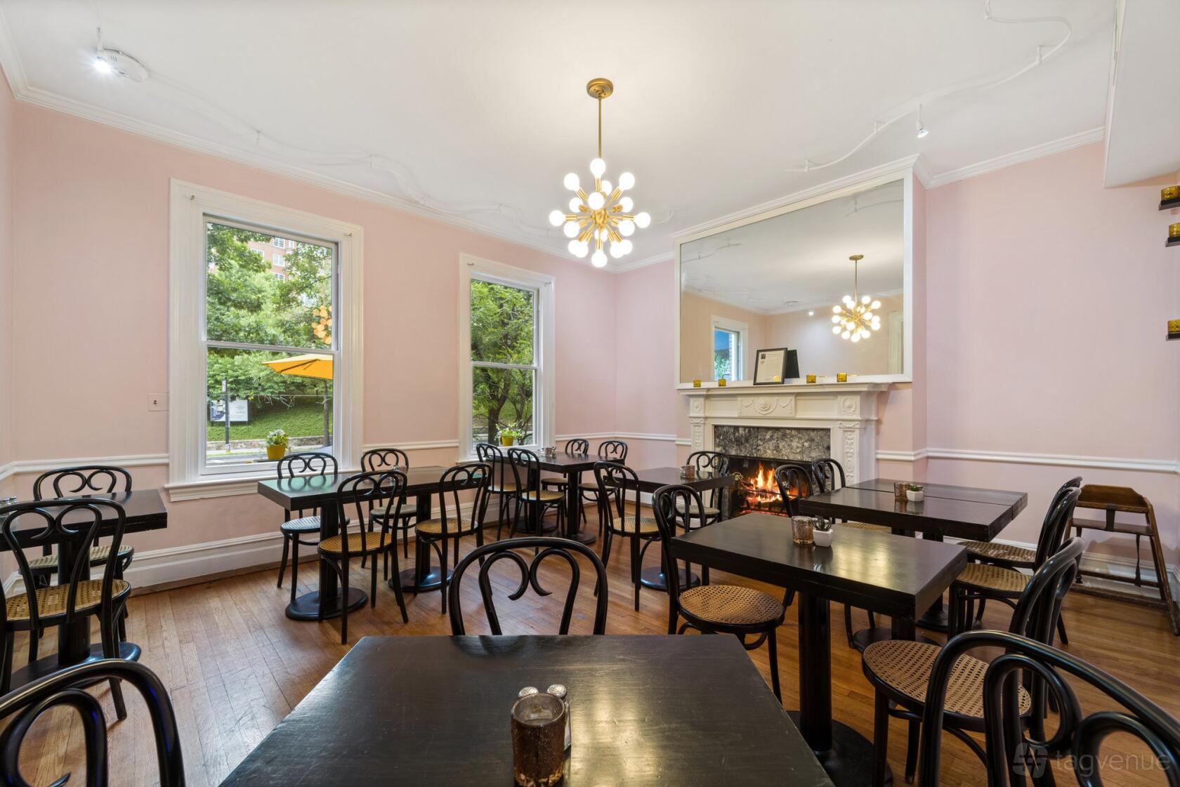 A dining room with black bistro chairs, pink walls, a decorative white fireplace, and large windows at Petit Monde Cafe.