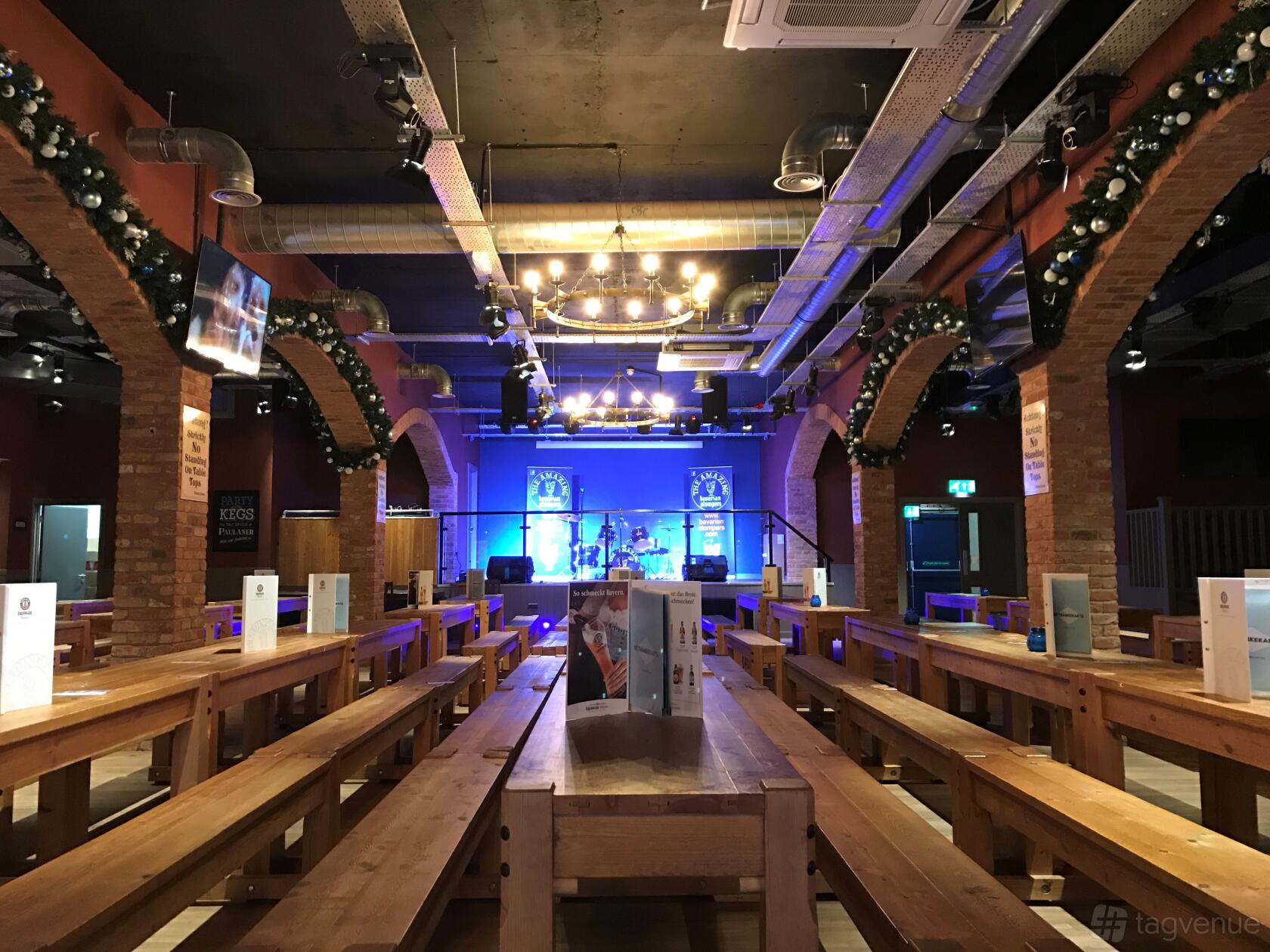 A bar with long wooden benches, exposed brick arches, and a stage under decorative lighting at The Bierkeller Birmingham.