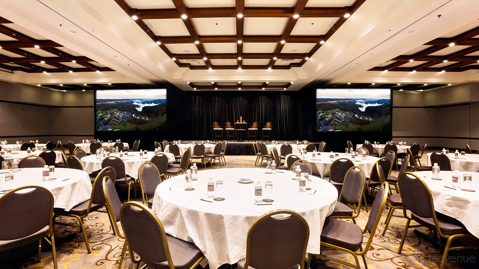 A hotel ballroom with round tables set with white linens, stage, and two large screens at Fairmont Resort Blue Mountains.