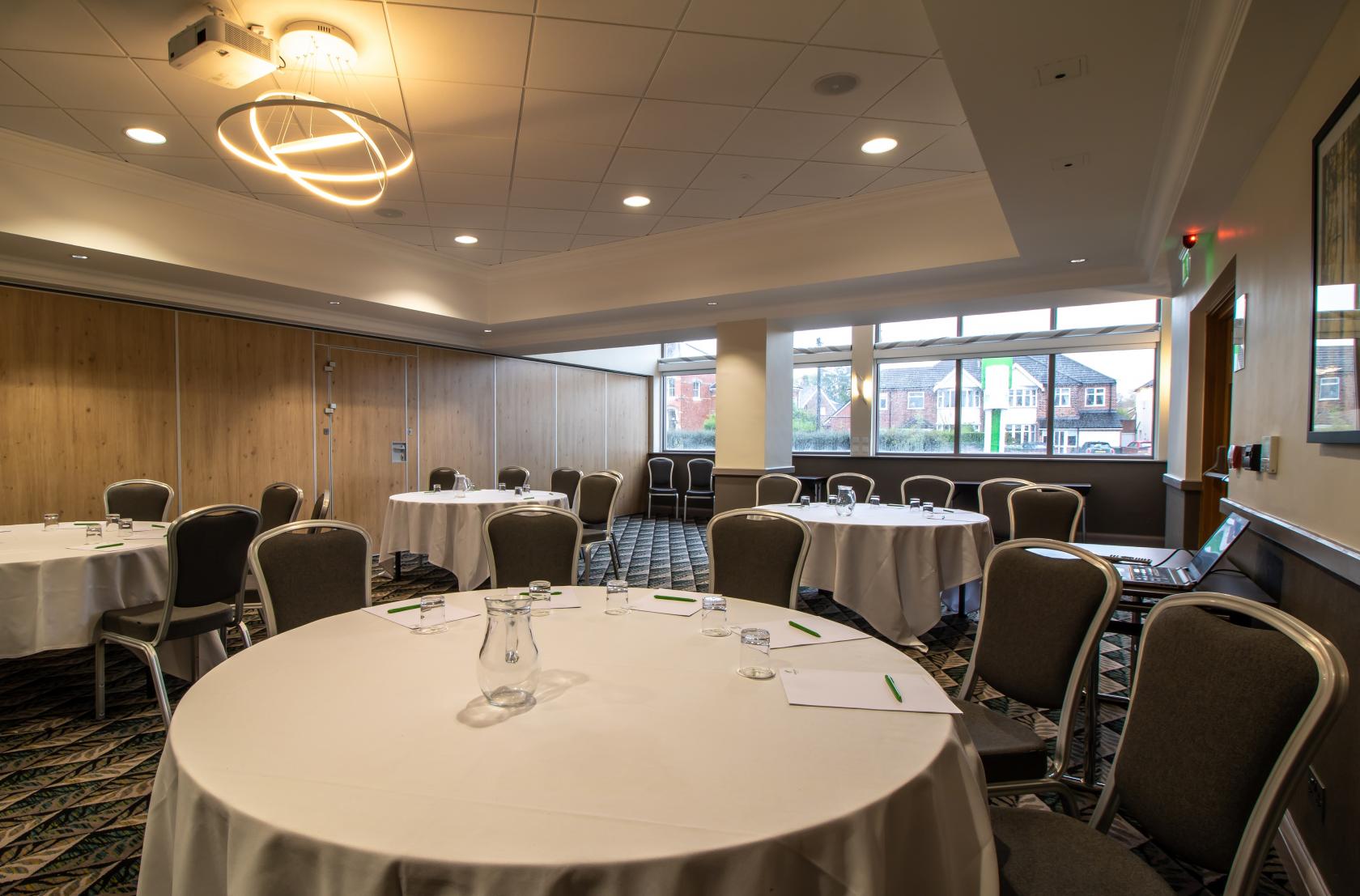 An event space with round tables draped in white linens, cushioned chairs, and large windows at Holiday Inn Leicester - Wigston.