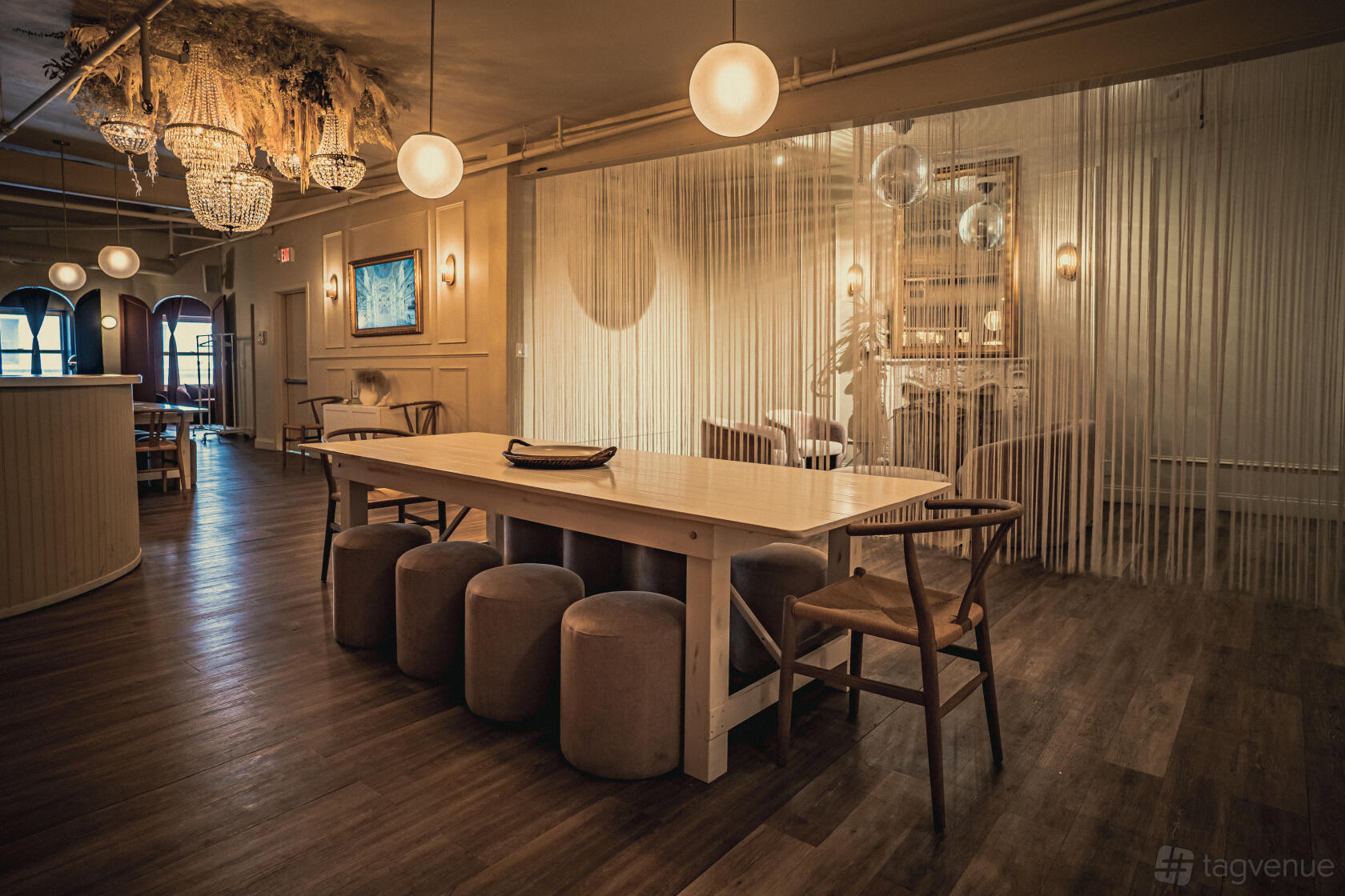 An event venue with a long wooden table, upholstered stools, warm pendant lighting, and sheer room dividers at Upscale Full Service Loft on 5th.
