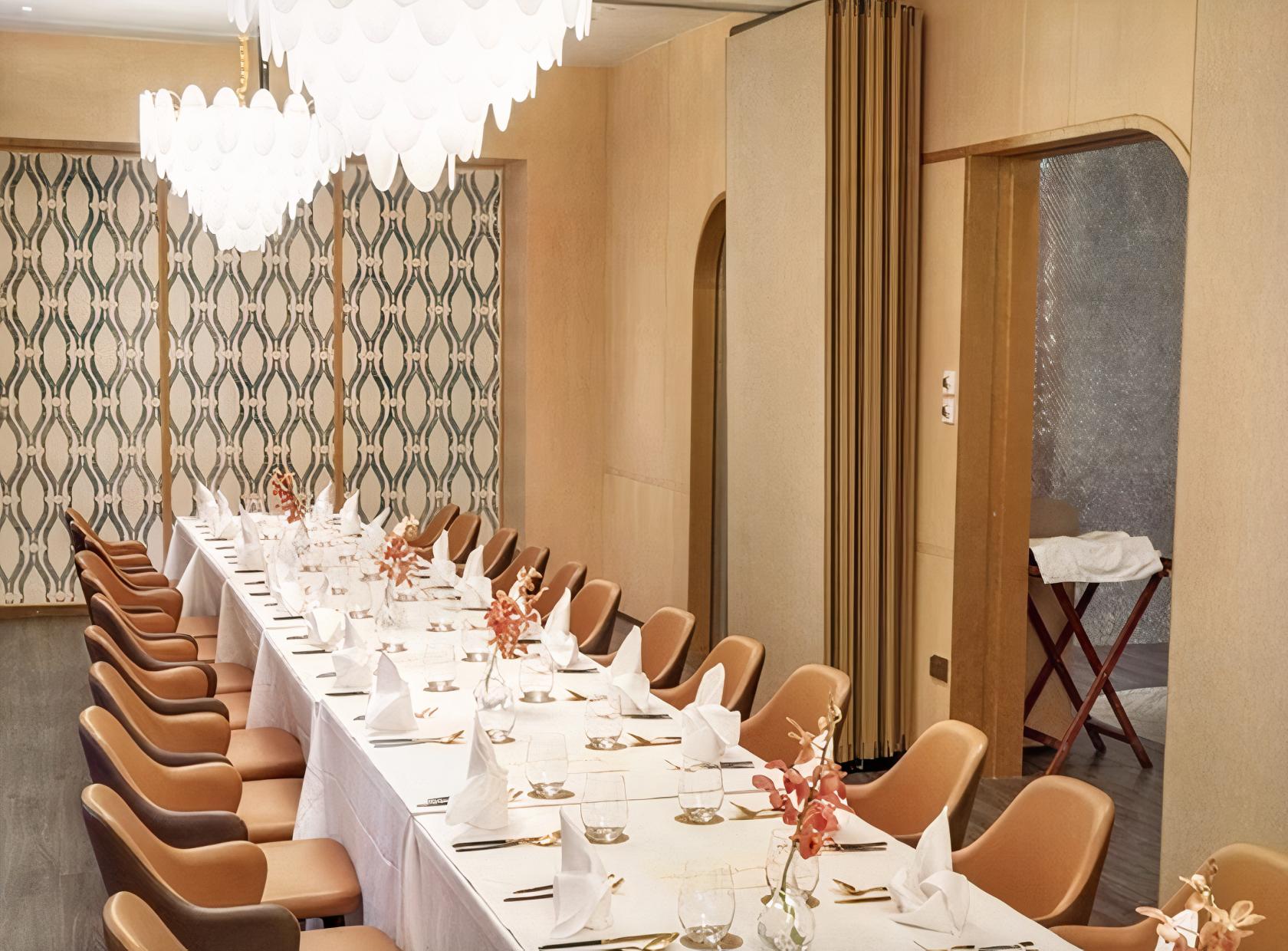 A dining room with a long banquet table, leather chairs, chandeliers, and patterned wall panels at SARAI FINE THAI.