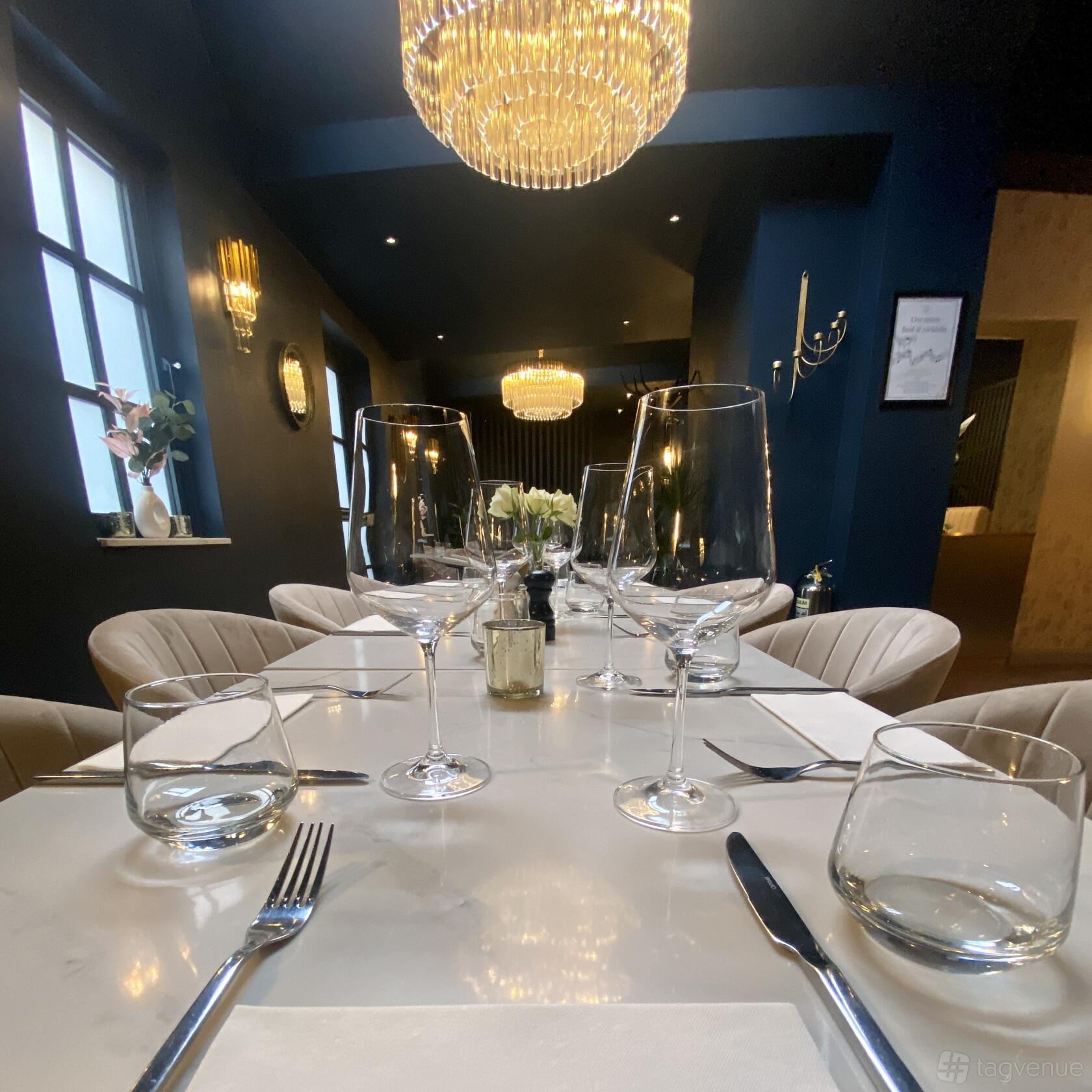 A restaurant dining room with a long white table, upholstered chairs, and chandeliers at Flame & Fire - Hammersmith.