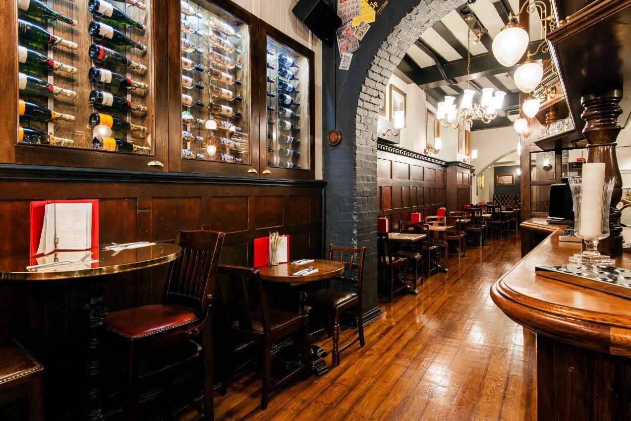 A basement bar with wooden floors, dark panelled walls, wine displays, and round tables at The Coal Hole.
