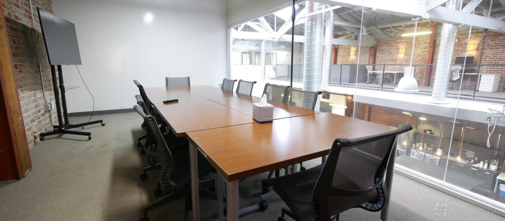 A coworking space with a long wooden meeting table, black mesh chairs, and floor-to-ceiling glass wall at Oakstop 2323 Broadway.