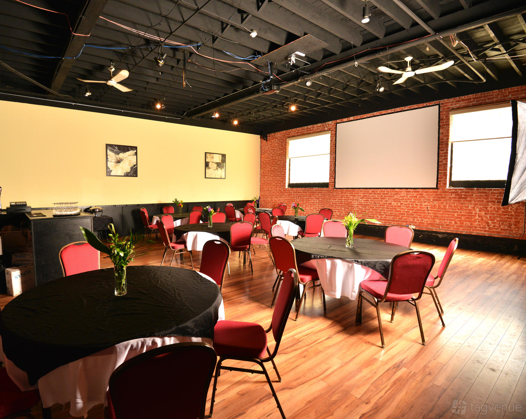 An event space with exposed brick walls, round tables with black linens, and red chairs at Maple Leaf House Grill & Lounge.