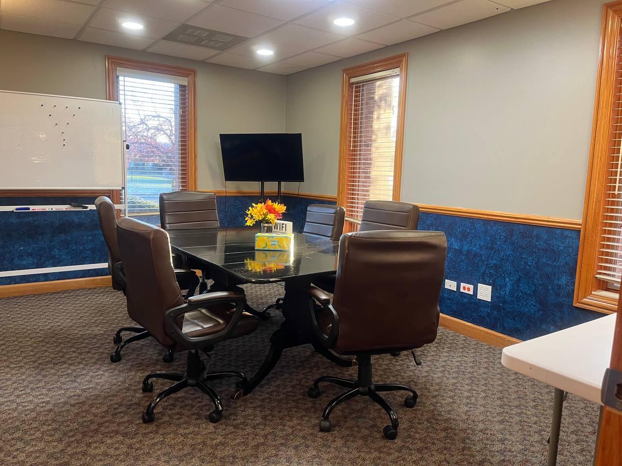 A meeting room with a glass conference table, black leather chairs, a wall-mounted TV, and windows at 7030 Centennial Center.