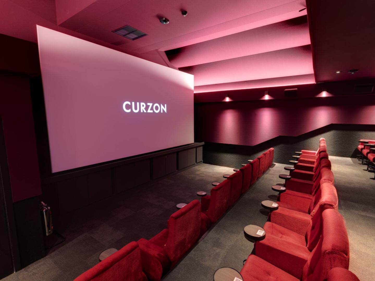 A cinema auditorium with red plush seats, individual tray tables, and a large projection screen at Curzon Camden.
