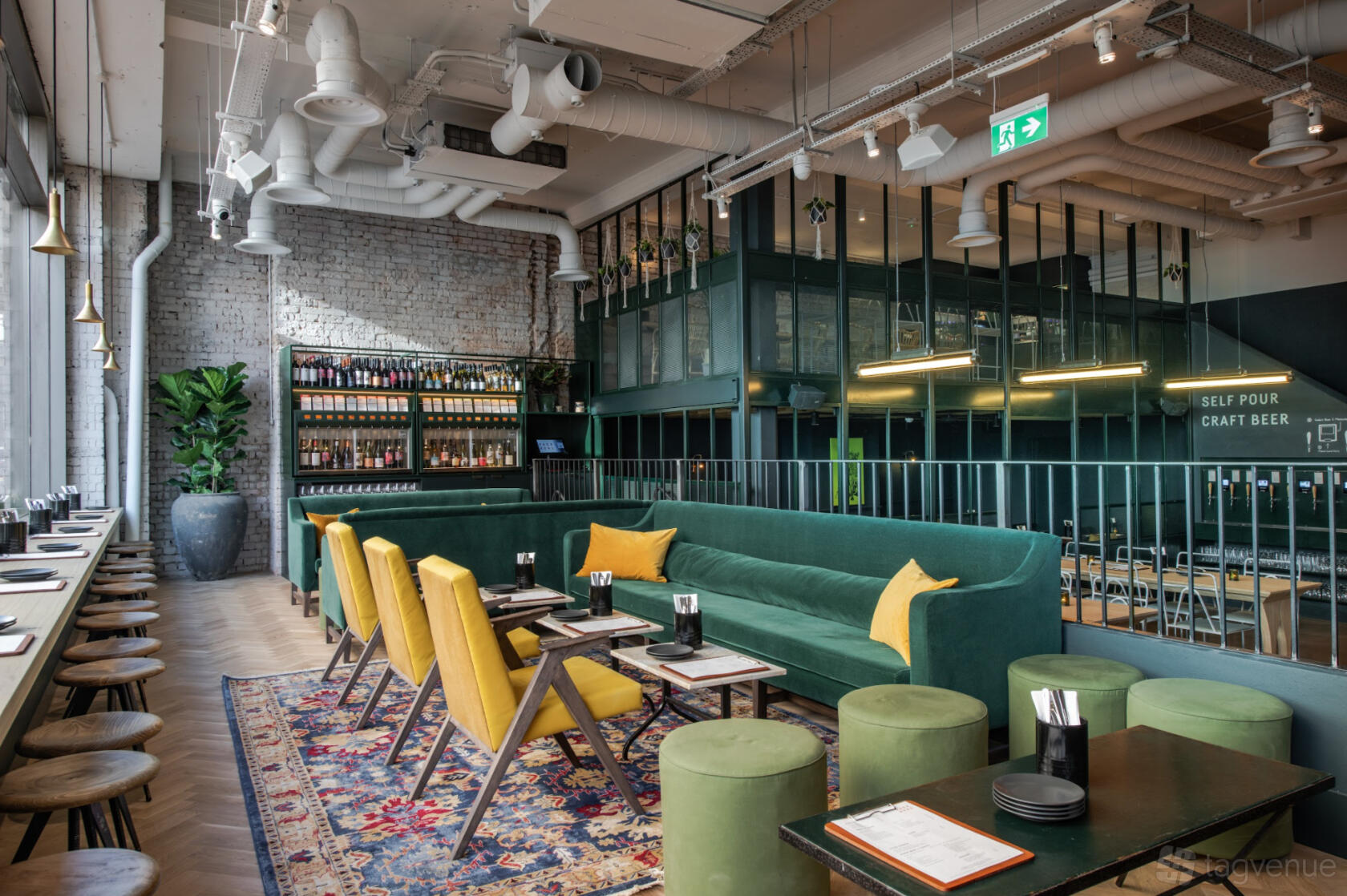An event space in a bar with a green velvet banquette, yellow chairs, exposed ductwork, and floor-to-ceiling windows at Vagabond Monument.