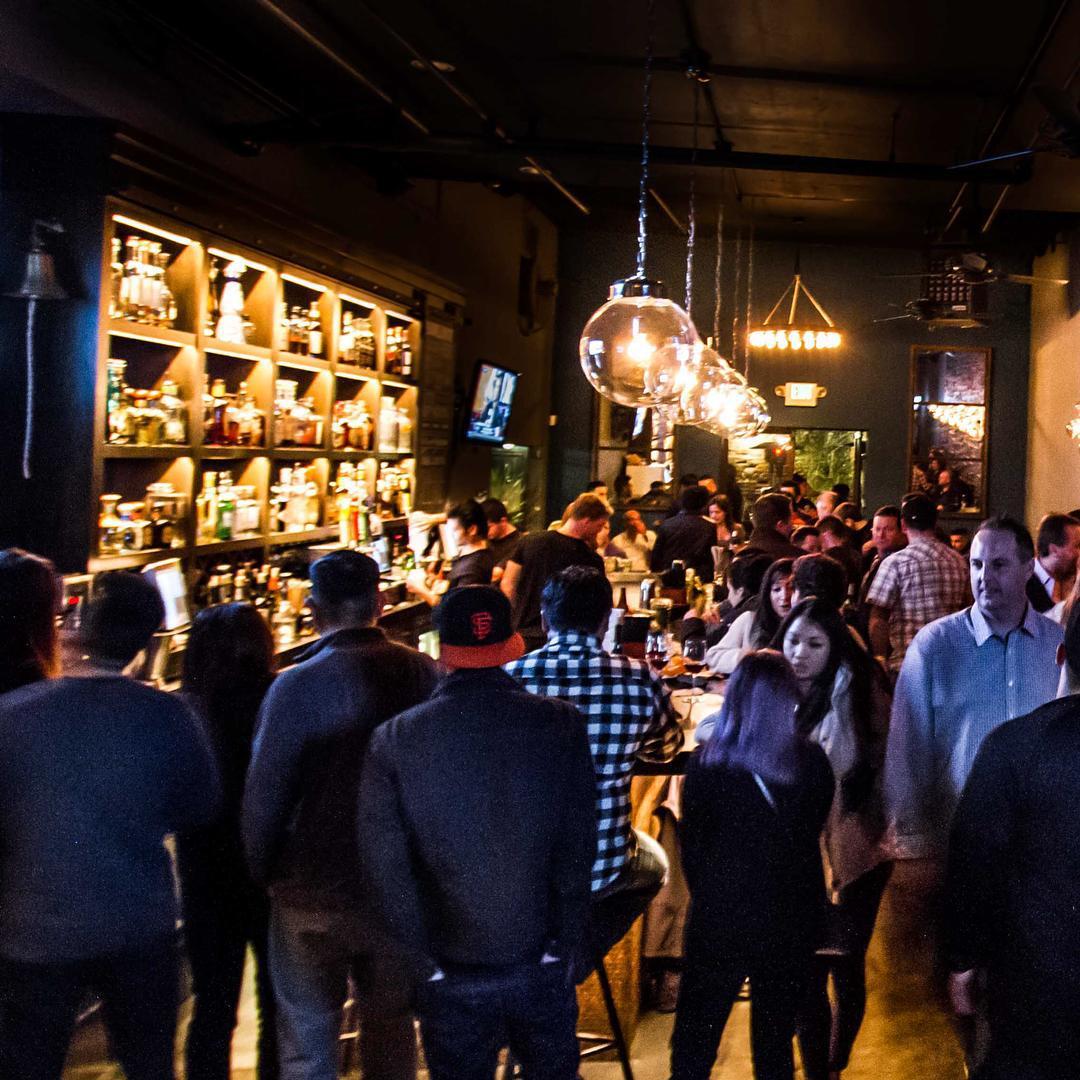 A bar with illuminated shelves stocked with liquor bottles, hanging globe lights, and a crowded counter at Barrelhouse.