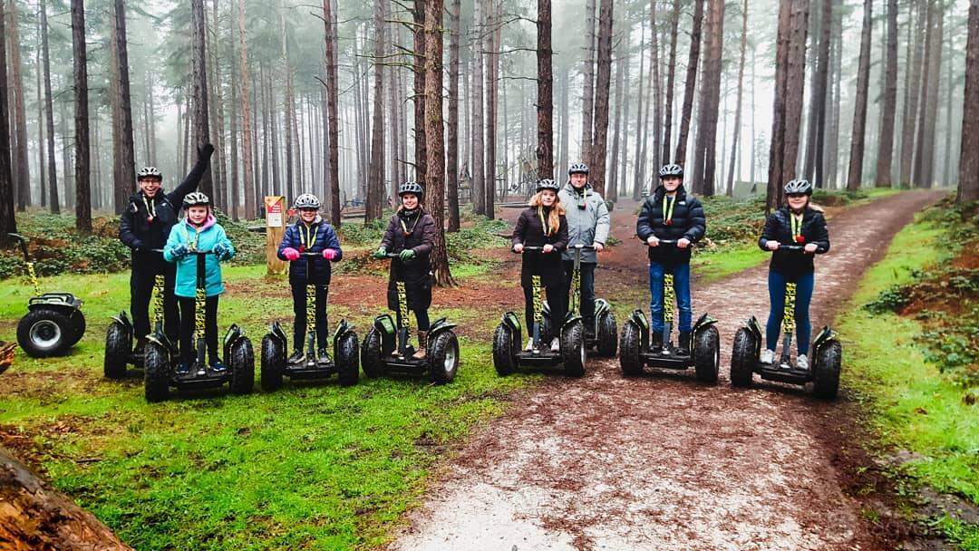 An outdoor event area with tall pine trees and a dirt path, featuring a group on Segways at Go Ape Sherwood Pines.