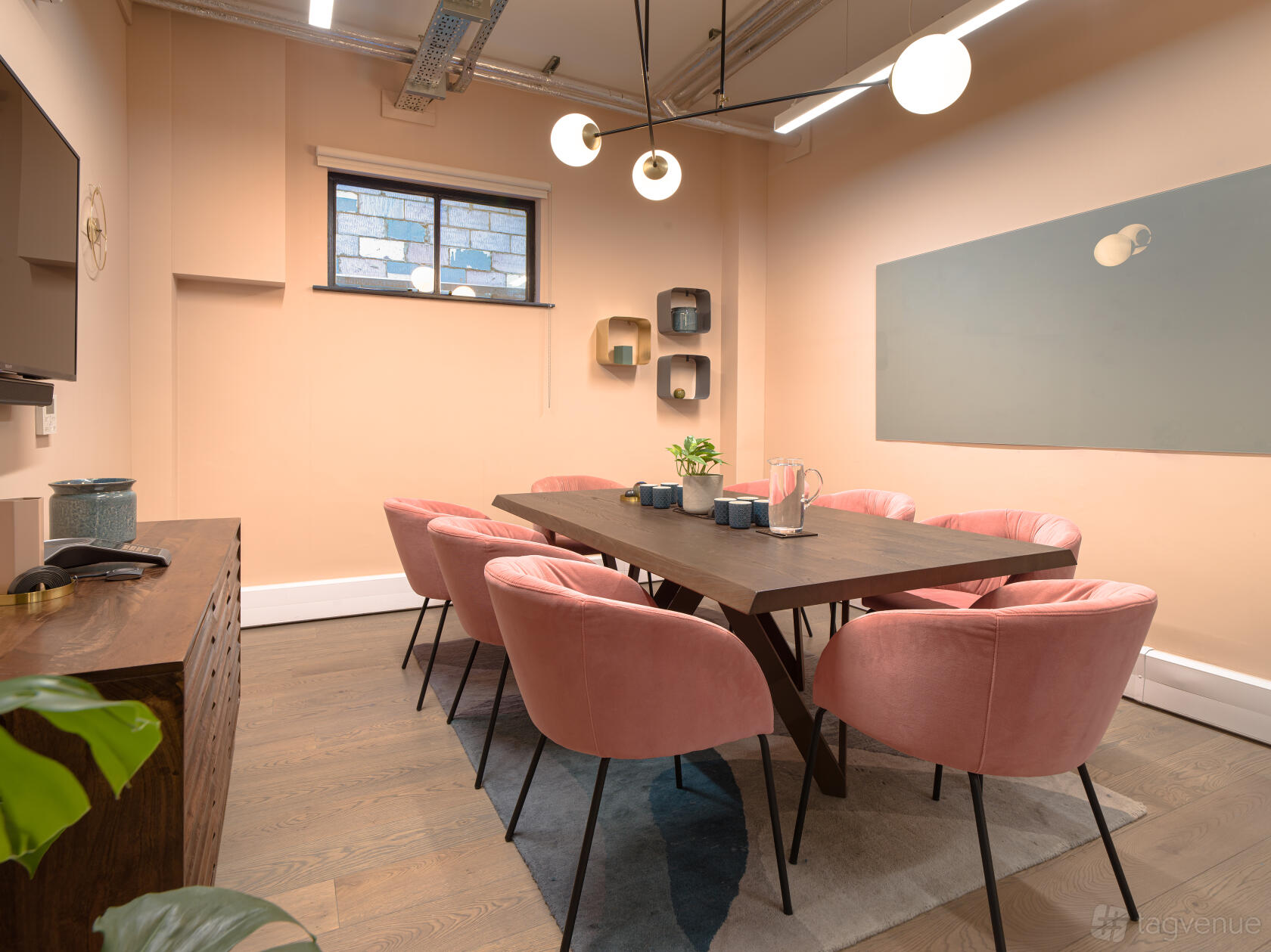A meeting room with a wooden table, pink velvet chairs, wall shelves, and globe pendant lighting at Uncommon Fulham.