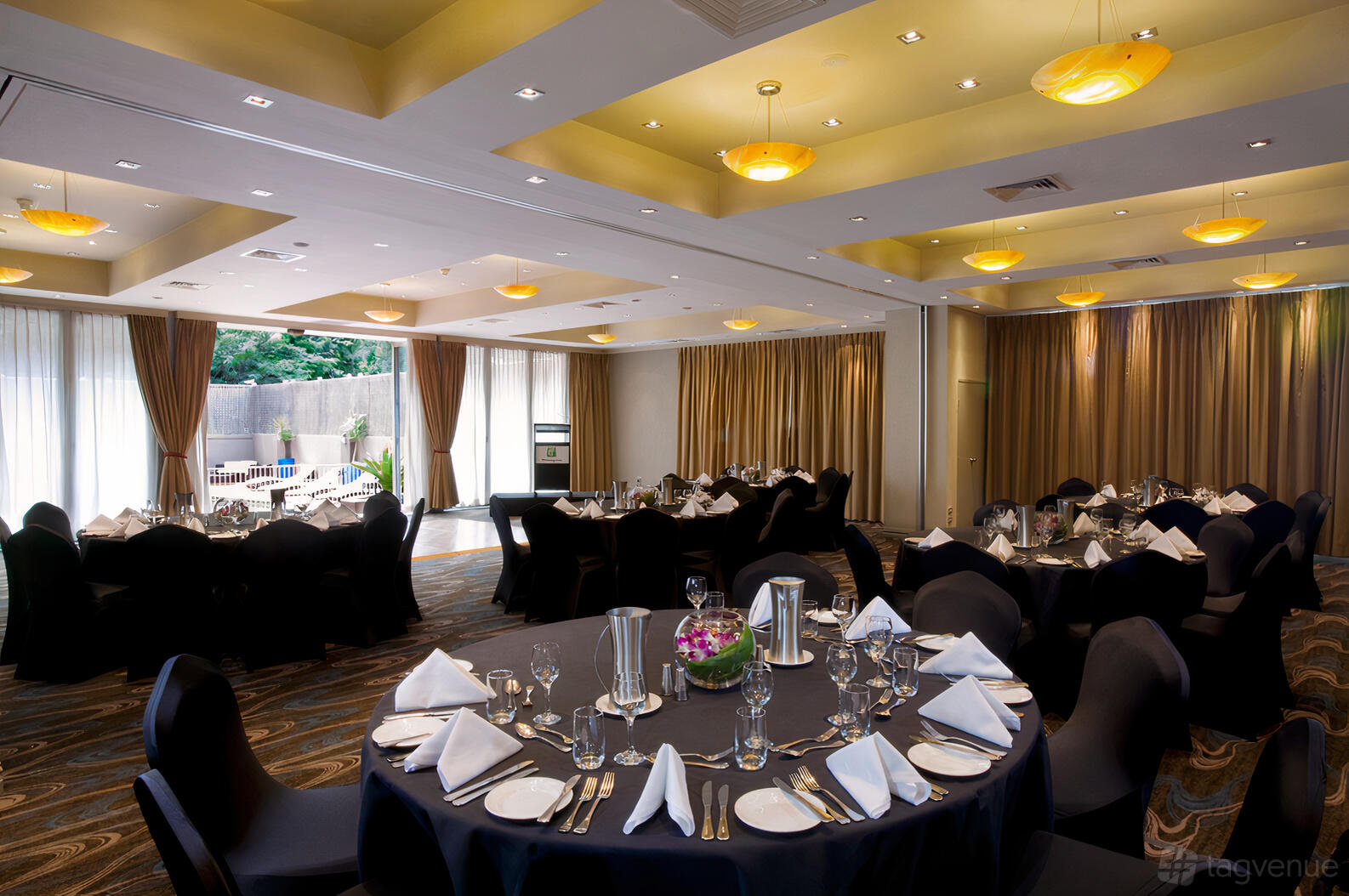 A hotel ballroom with round tables, black chair covers, white napkins, and floor-to-ceiling windows at Holiday Inn Parramatta.