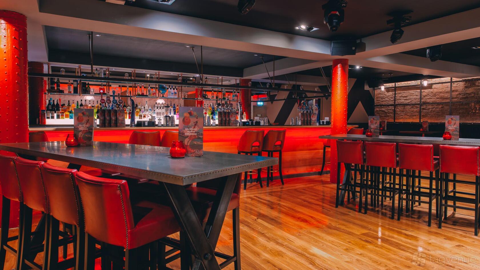 An event space in a bar with a long illuminated bar, red leather stools, and high tables at Revolution Mitchell Street.