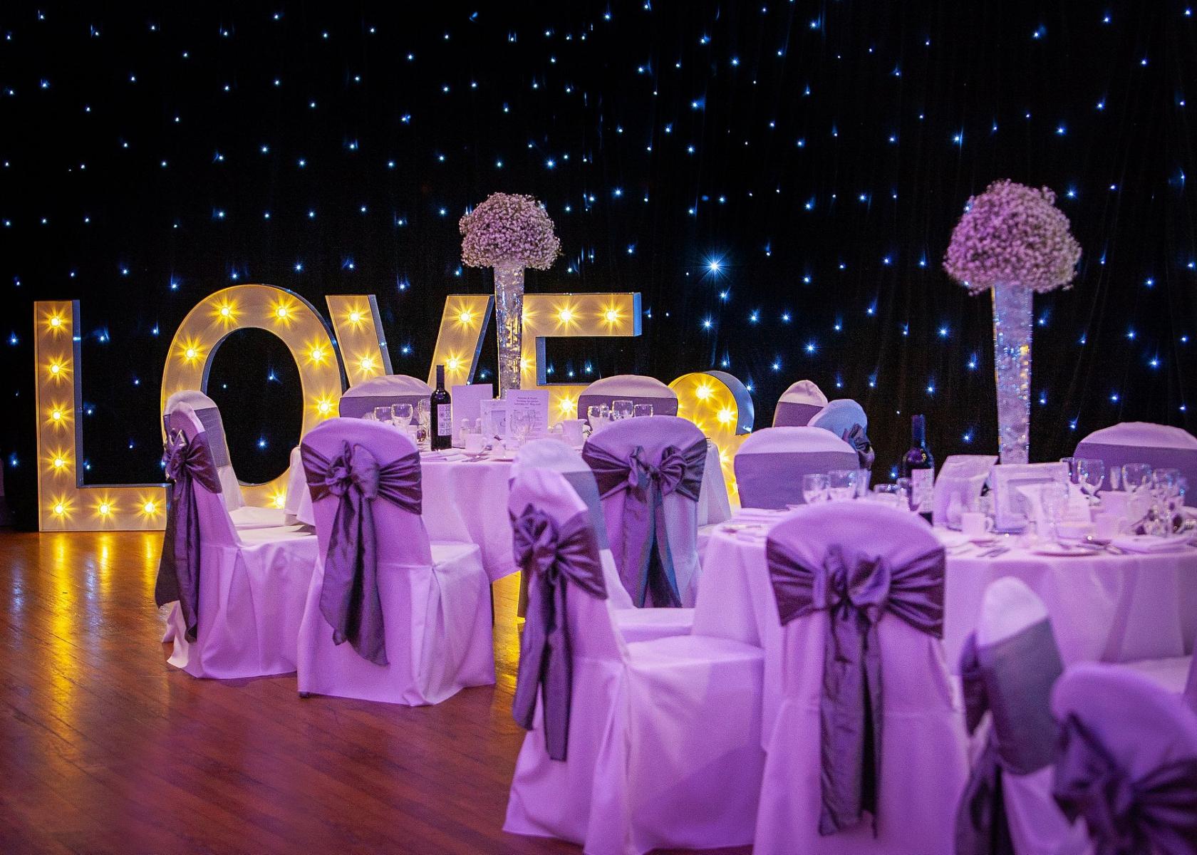 An event venue with round tables draped in white linen and purple sashes, large illuminated 'LOVE' letters, and starry backdrop at Edinburgh Corn Exchange.