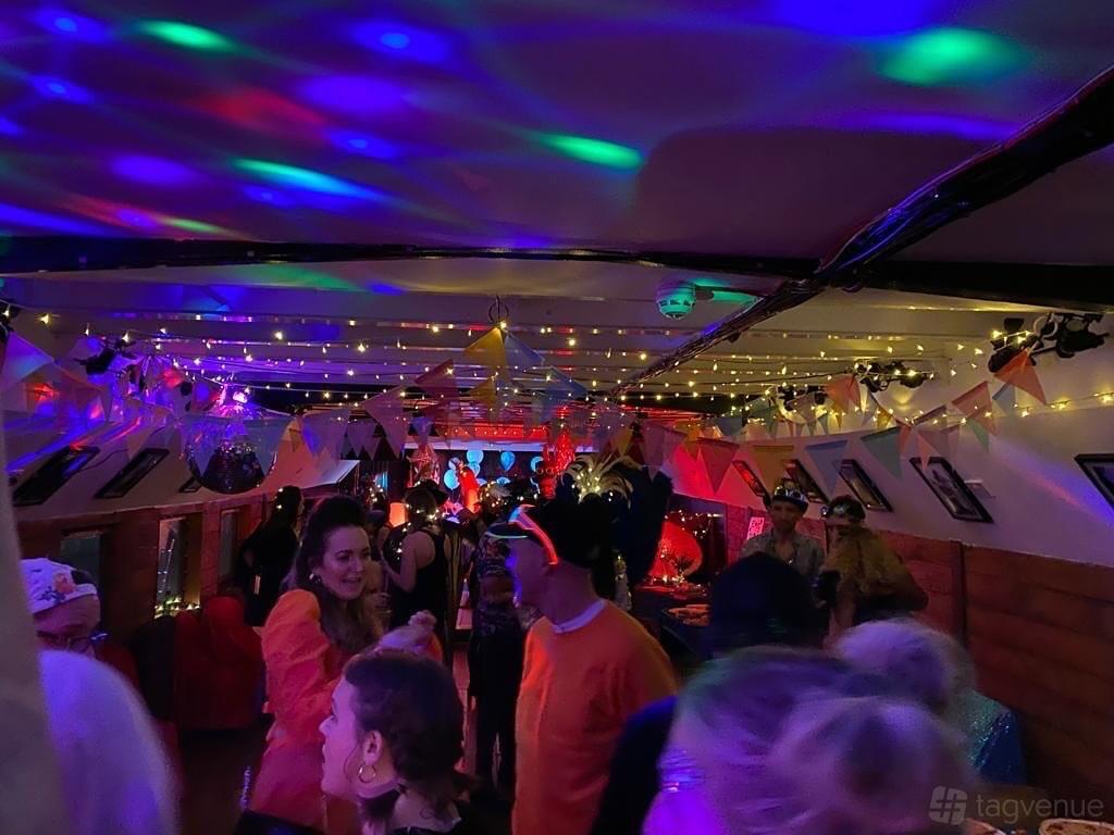 An event space on a boat with colorful disco lights, party decorations, and guests dancing on the Lower Deck at Battersea Barge.