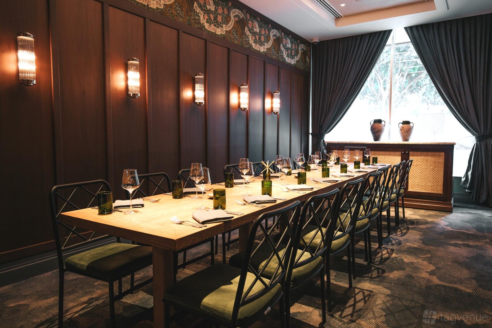A dining room with a long wooden table, upholstered chairs, wall sconces, and floor-to-ceiling curtains at Ovolo The Valley.