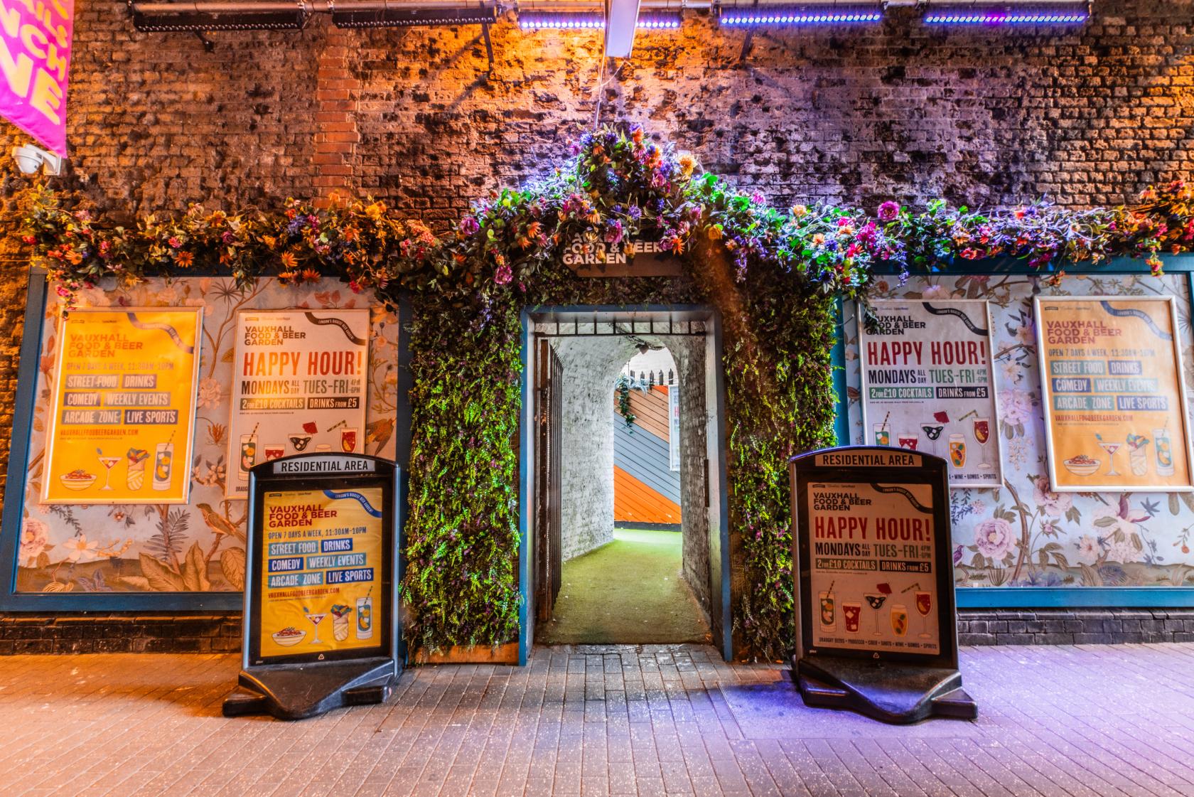 An open-air venue with a floral archway entrance, exposed brick, and colorful poster signage at Vauxhall Food and Beer Garden.