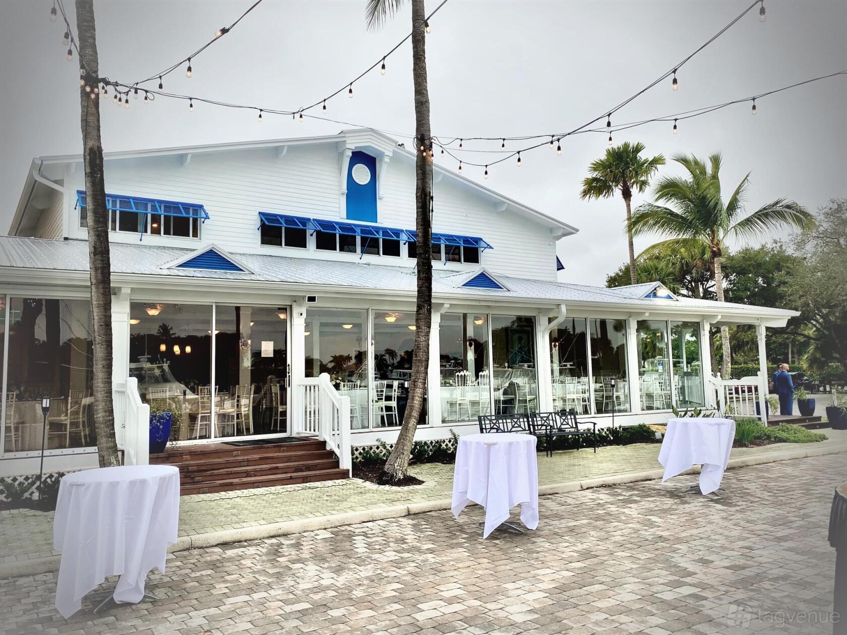 An event venue with a white building, string lights, palm trees, and cocktail tables at Out of the Blue Celebrations.