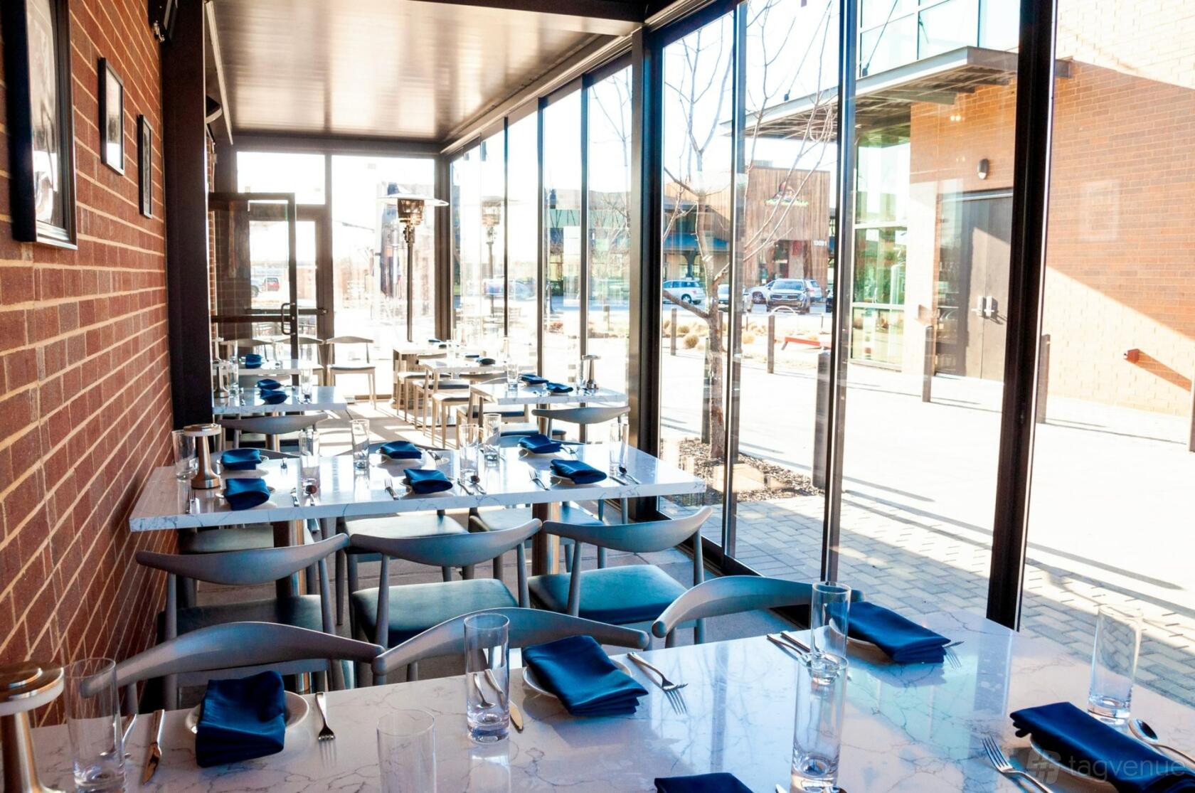A terrace with floor-to-ceiling glass walls, brick accent wall, and marble tables set with blue napkins at Roots Southern Table.
