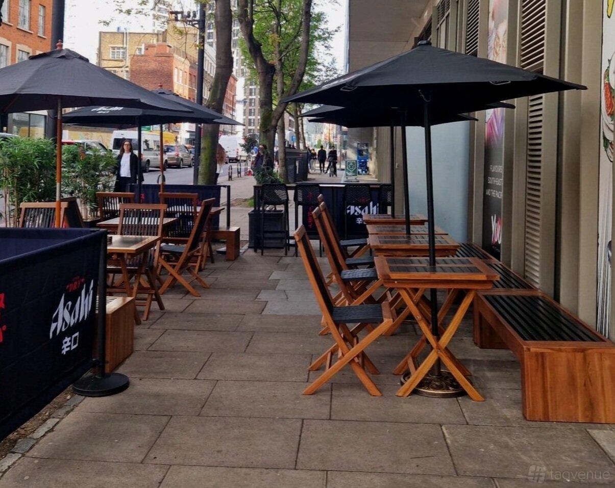 An outdoor terrace with wooden tables, folding chairs, and black umbrellas at YUU Spitalfields.