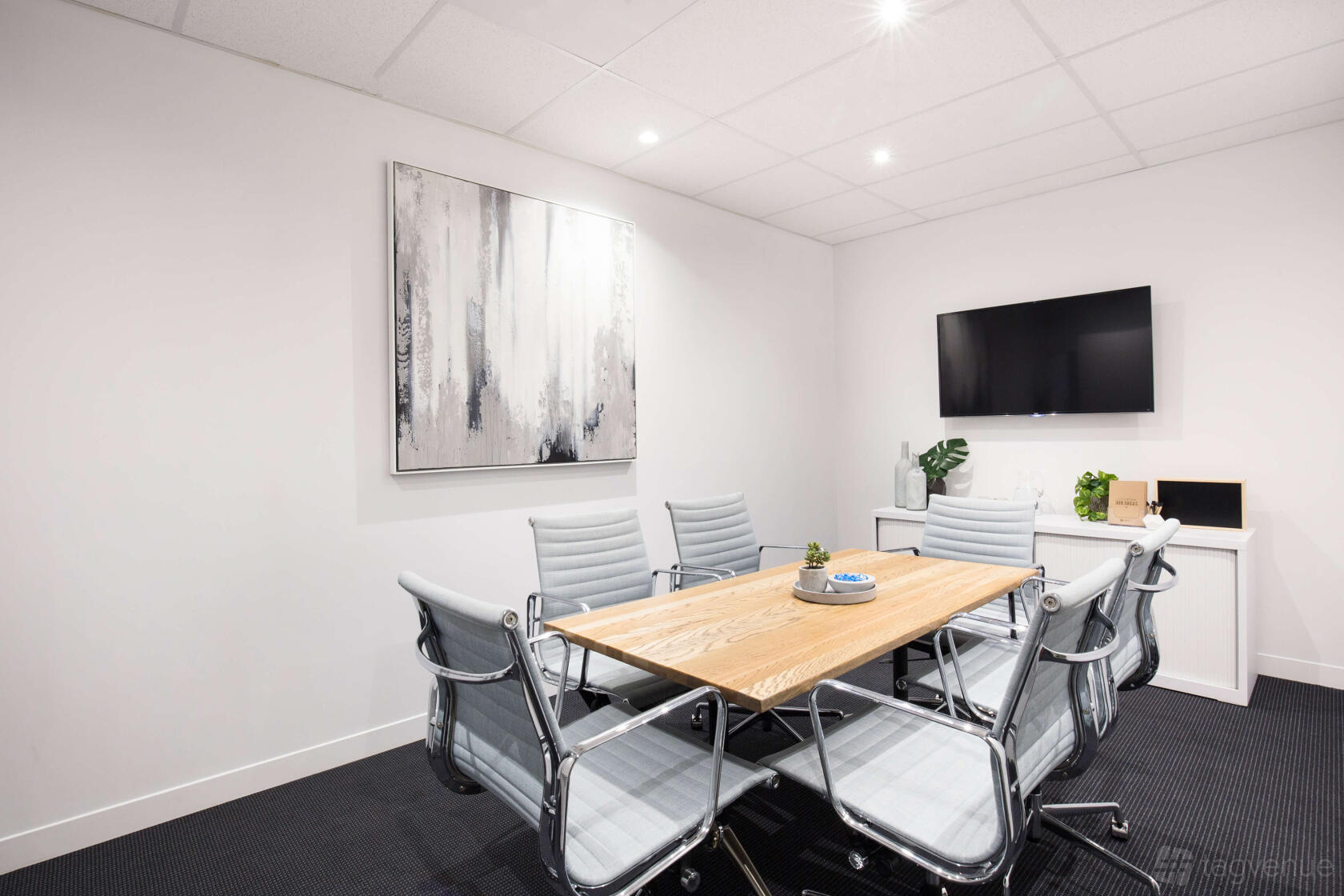 A meeting room with a wooden conference table, six gray chairs, wall art, and a mounted TV at Peninsula On The Bay.