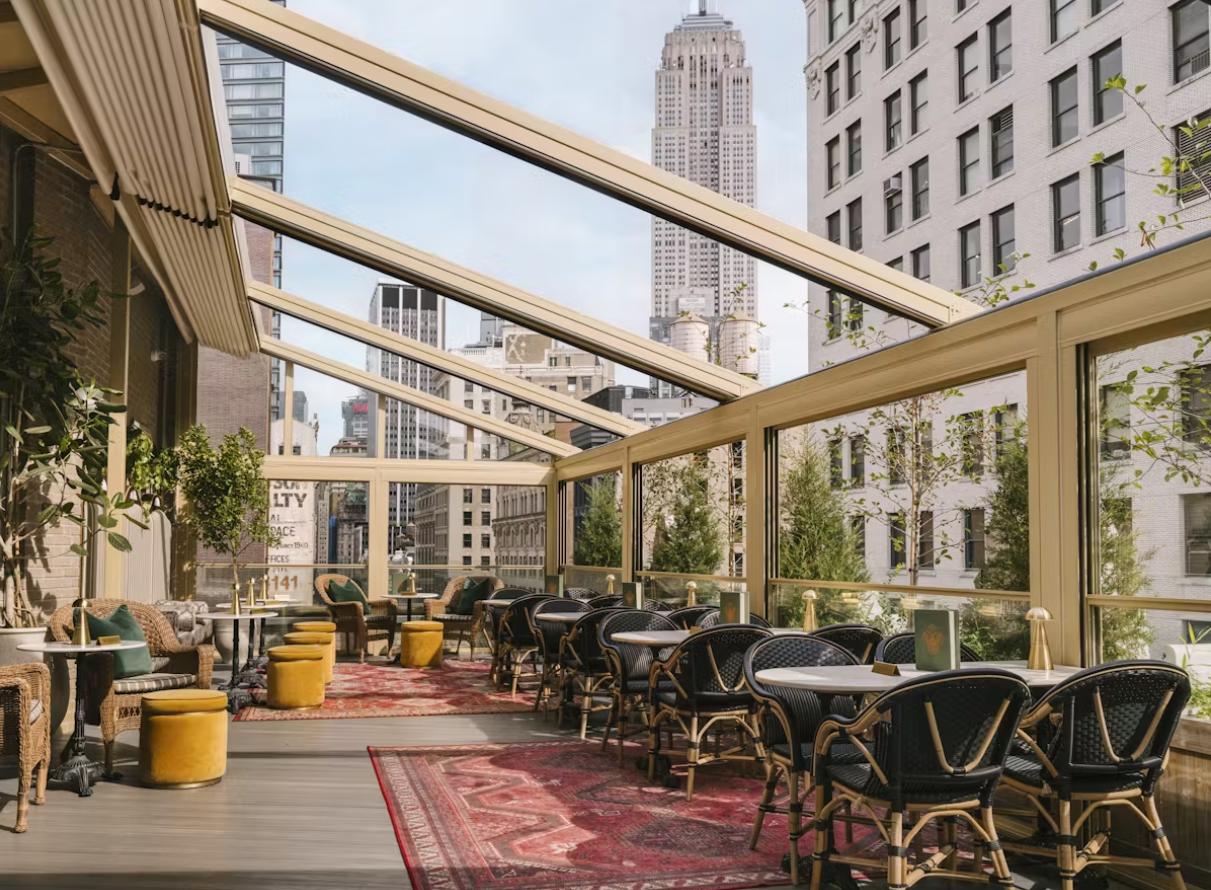 A terrace with glass walls, bistro seating, patterned rugs, and city views at Apotheke NoMad.