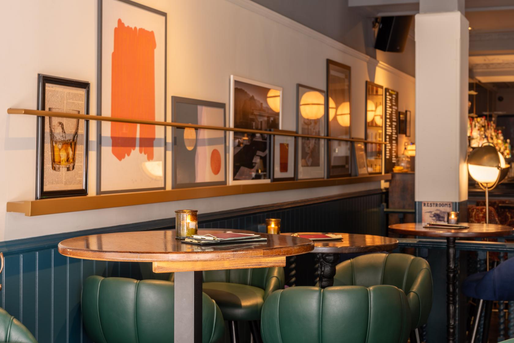 An event space in a cocktail bar with marble-topped tables, green leather chairs, and framed art at LOCKES Bar Covent Garden.