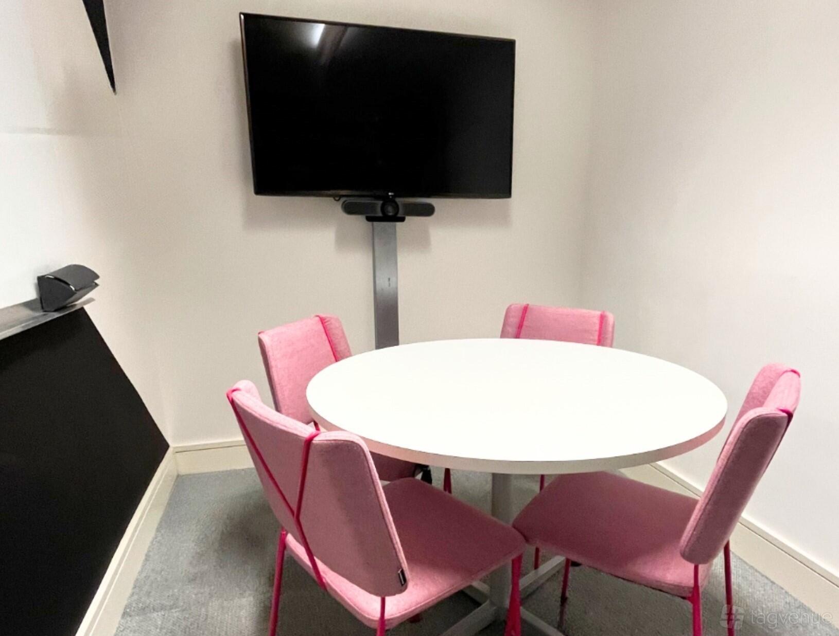 A coworking space with a round table, pink chairs, and a wall-mounted display screen at Huckletree D2.