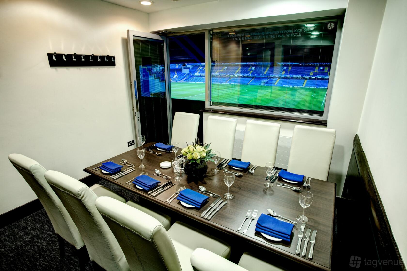 A function room with a set dining table, white leather chairs, and a large window overlooking the pitch at Chelsea Football Club.