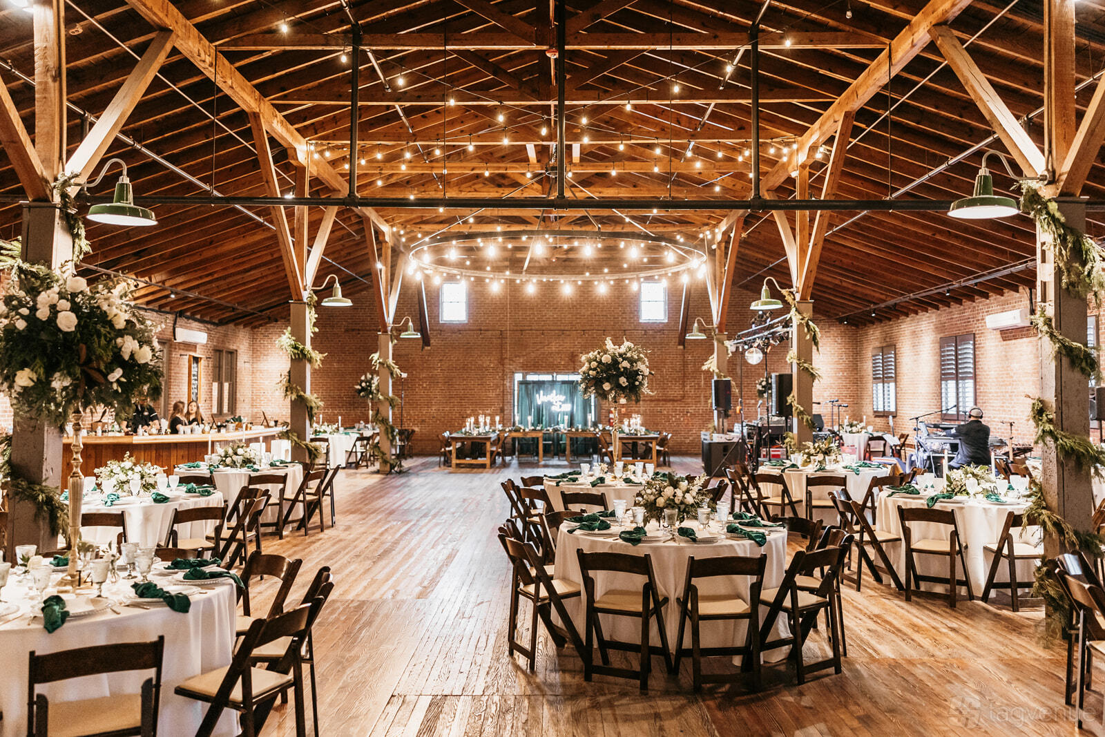 An event venue with exposed wooden beams, brick walls, round tables with white linens, and string lights at The Chair Factory Venue.