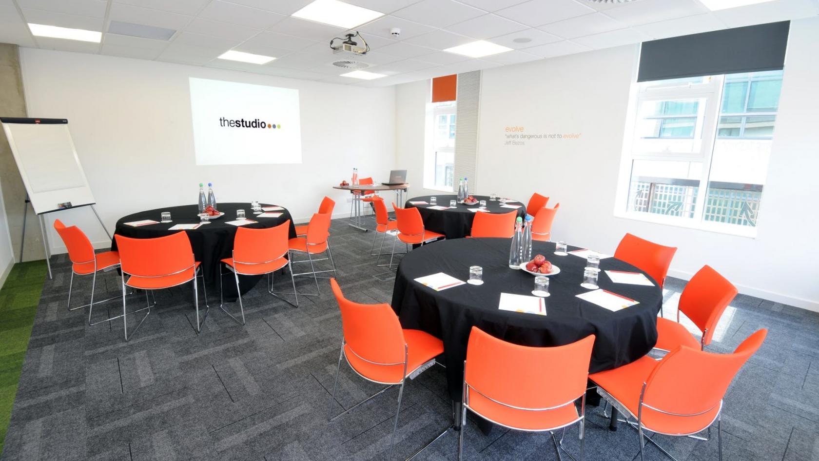 A function room with round tables, orange chairs, a projector screen, and large windows at The Studio Manchester.