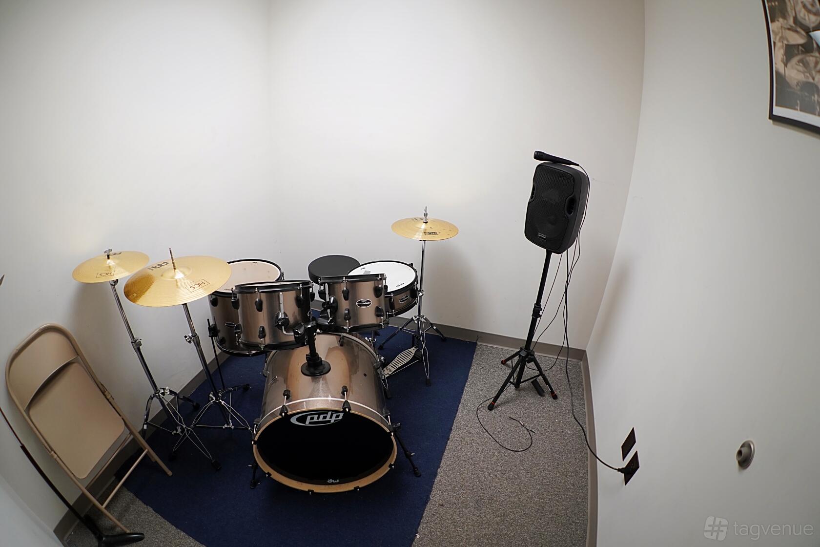 A rehearsal room with a drum kit, foldable chair, and speaker on a carpeted corner at Rehearse Live.