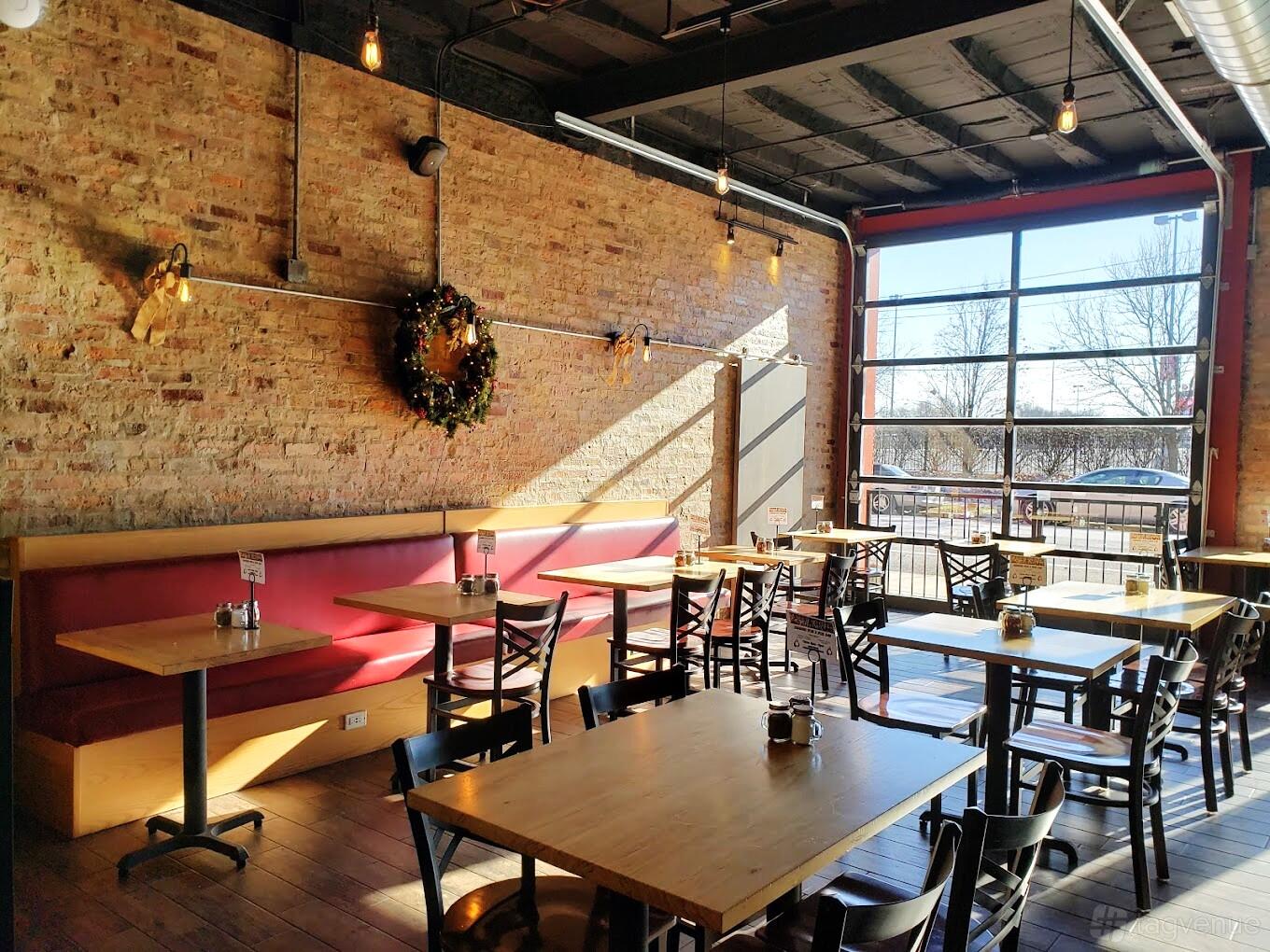 A restaurant with exposed brick walls, large garage-style windows, and wooden tables at Stix n Brix Wood Fired Pizza.