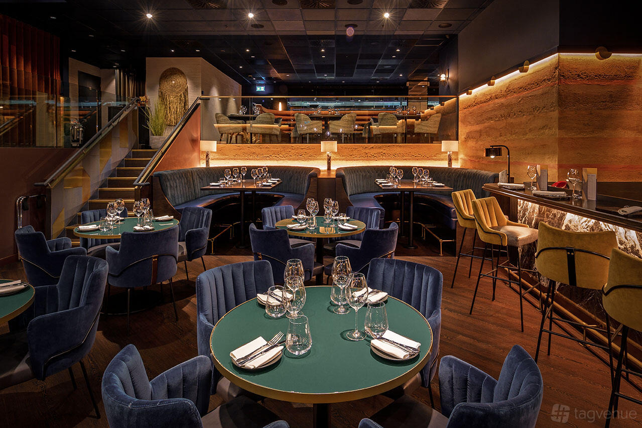 A restaurant with velvet blue chairs, round tables set with glassware, and warm recessed lighting at Gaucho Charlotte Street.