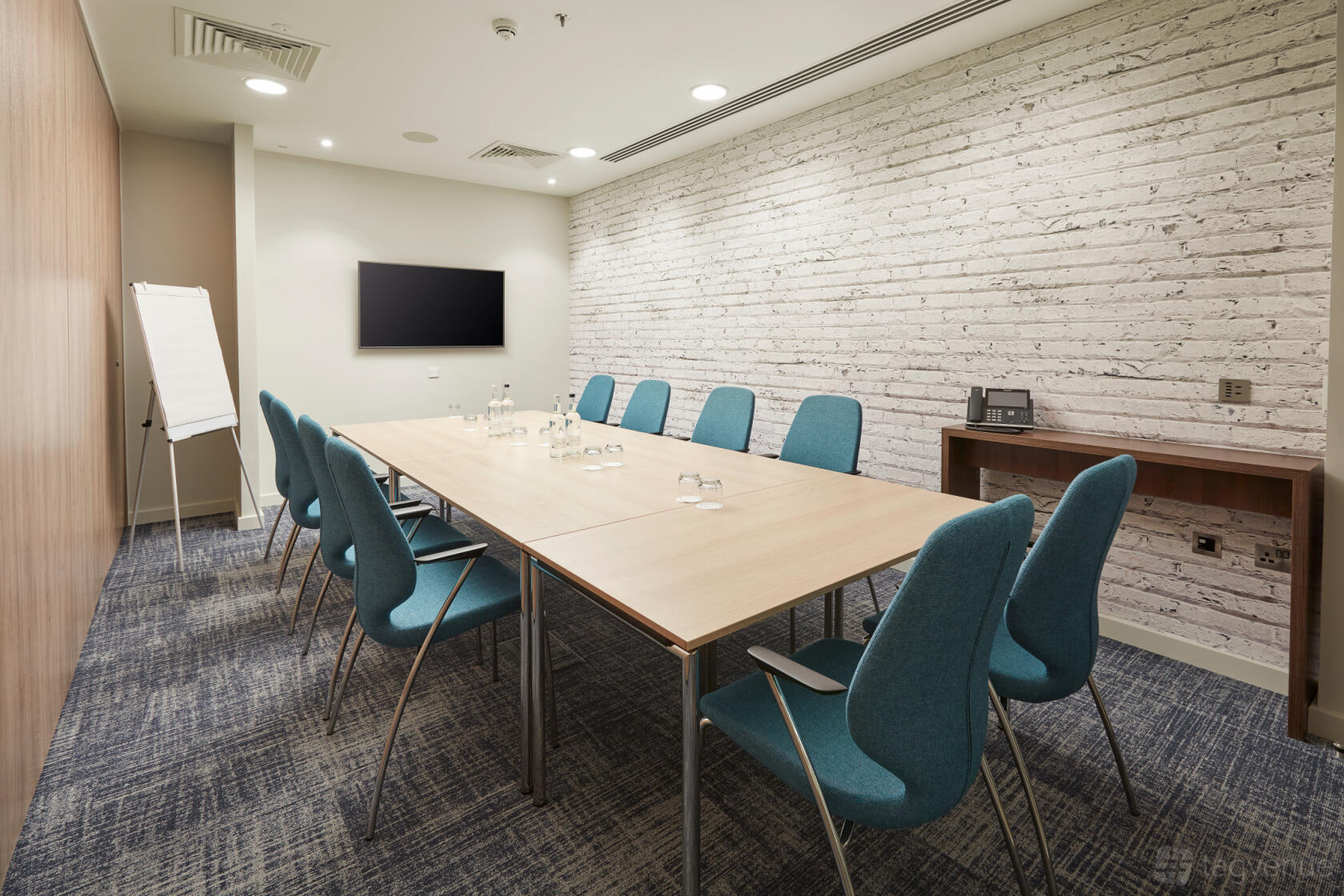 A meeting room with a long table, teal chairs, a white brick accent wall, and a mounted TV at Marlin Waterloo.
