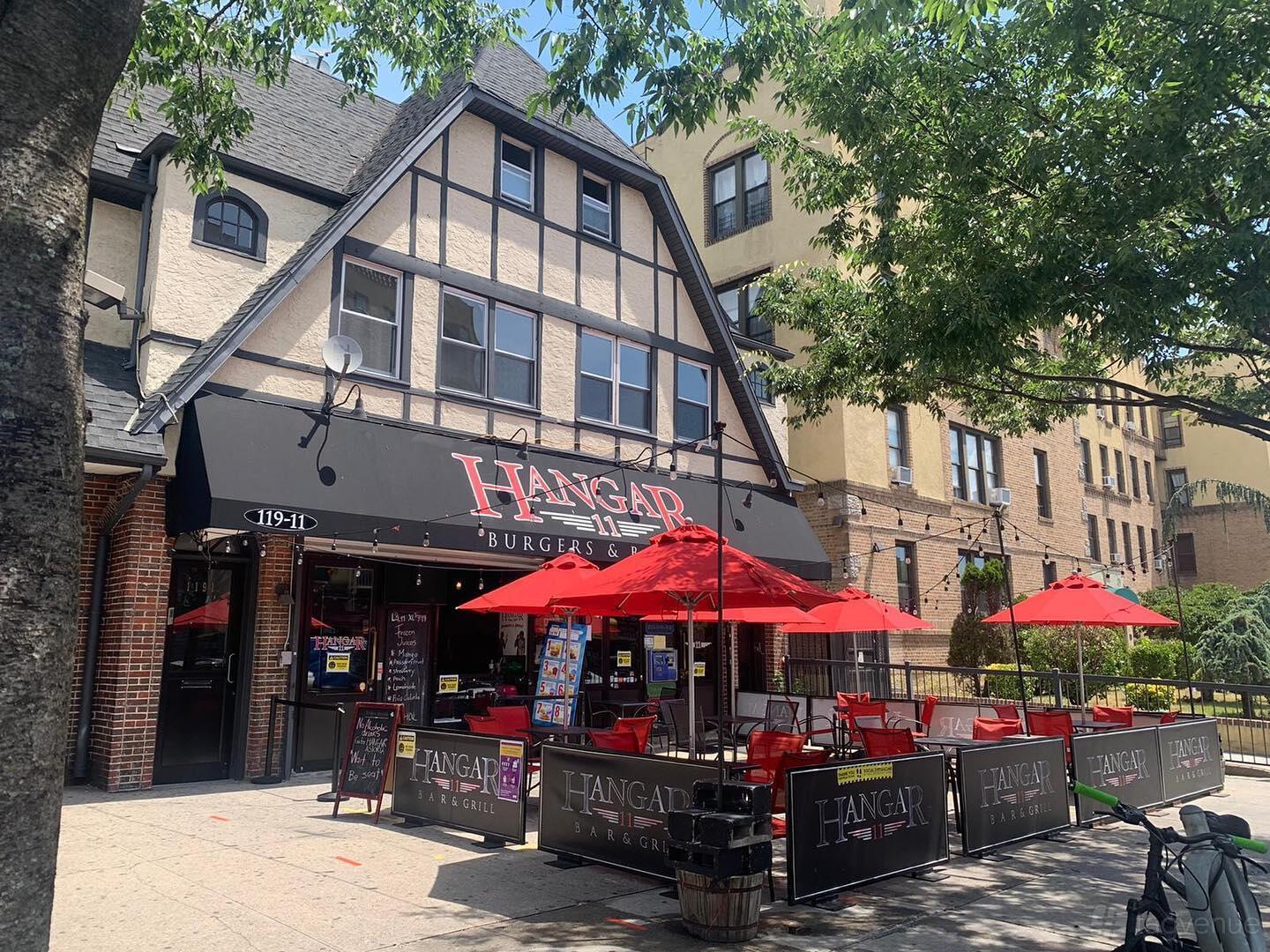 A bar with Tudor-style facade, outdoor patio seating, and red umbrellas at Hangar 11 Burgers & Brews.